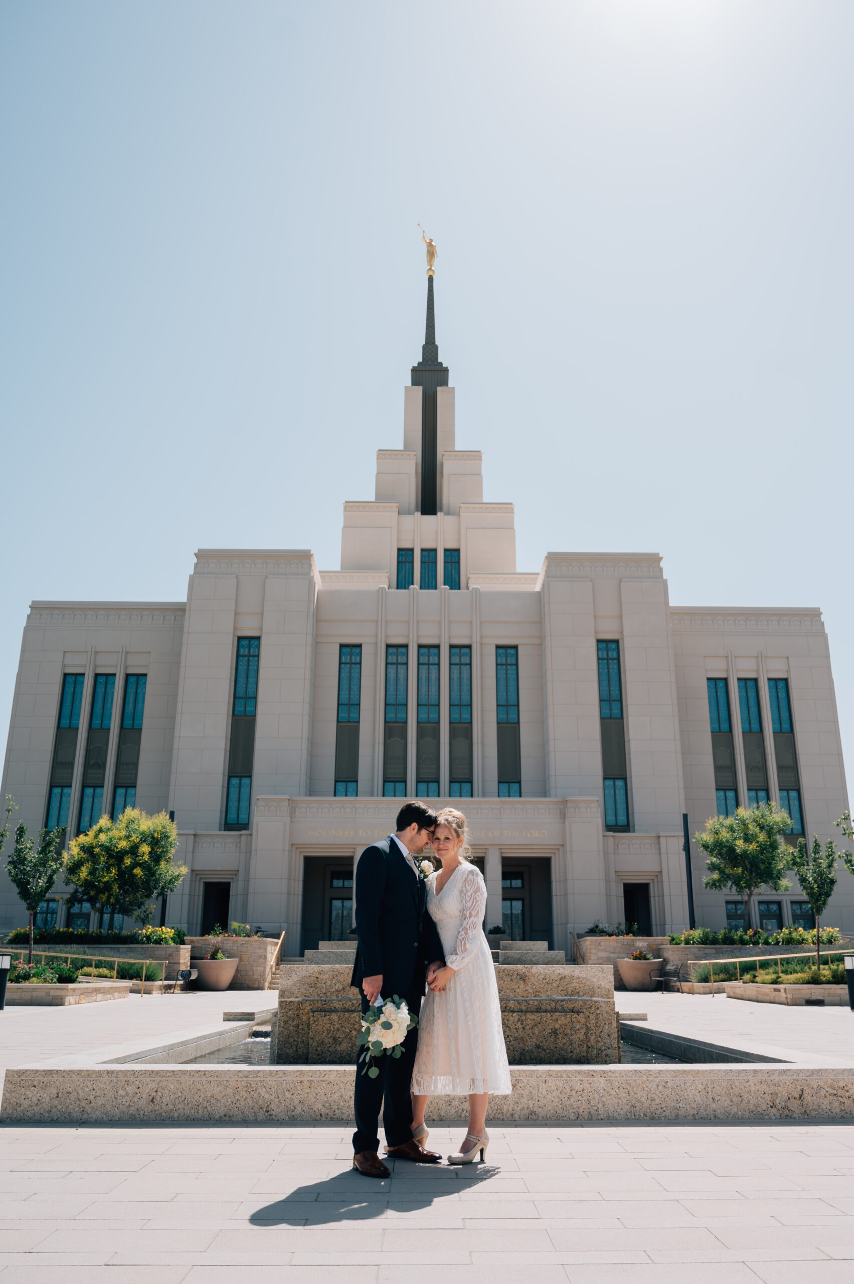 Groom leans in on his cute bride's temple for a tender moment while holding her flowers. She holds his hand with both of his.