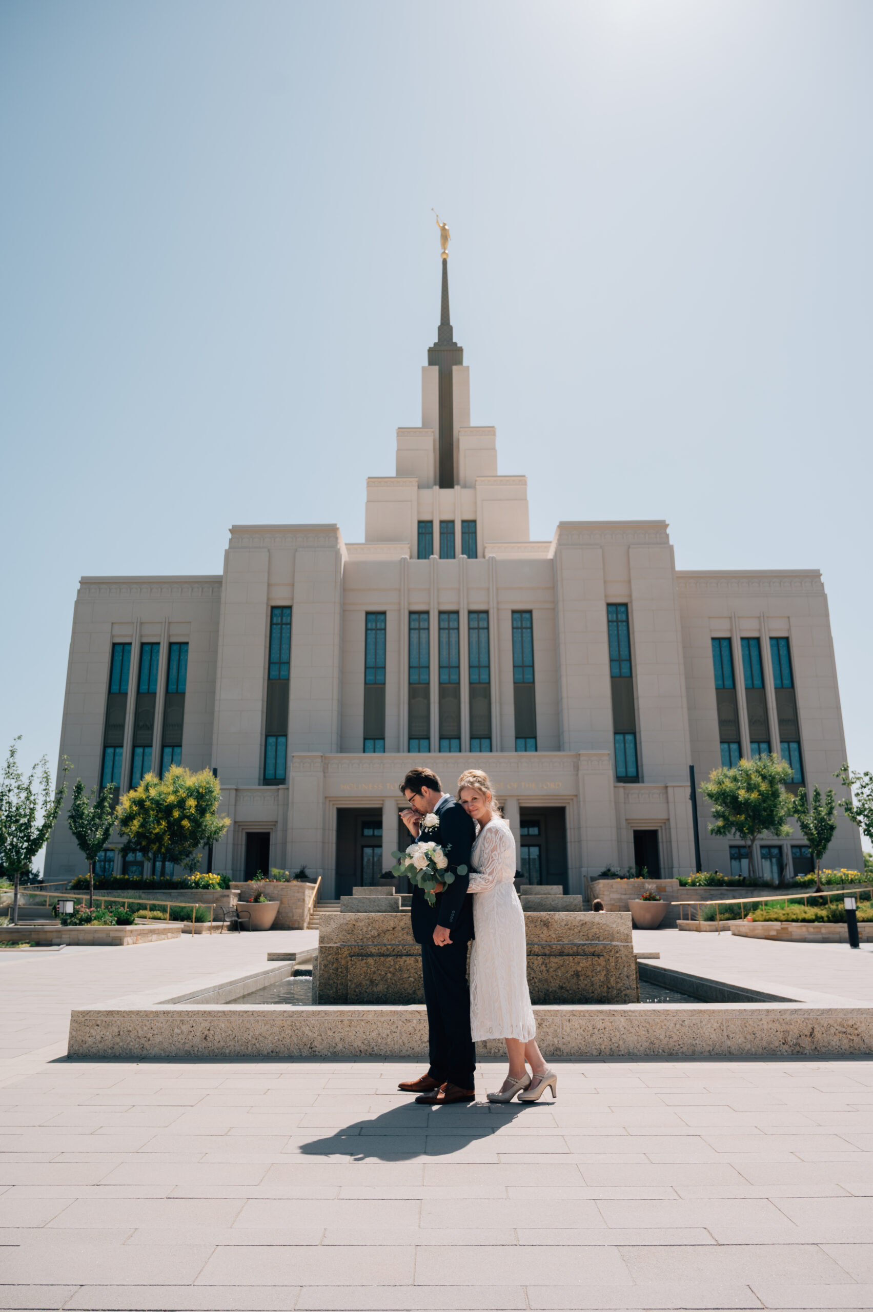 Groom kisses his brides hand in front of the LDS temple in Saratoga Springs Ut while she cuddles up from behind him.
