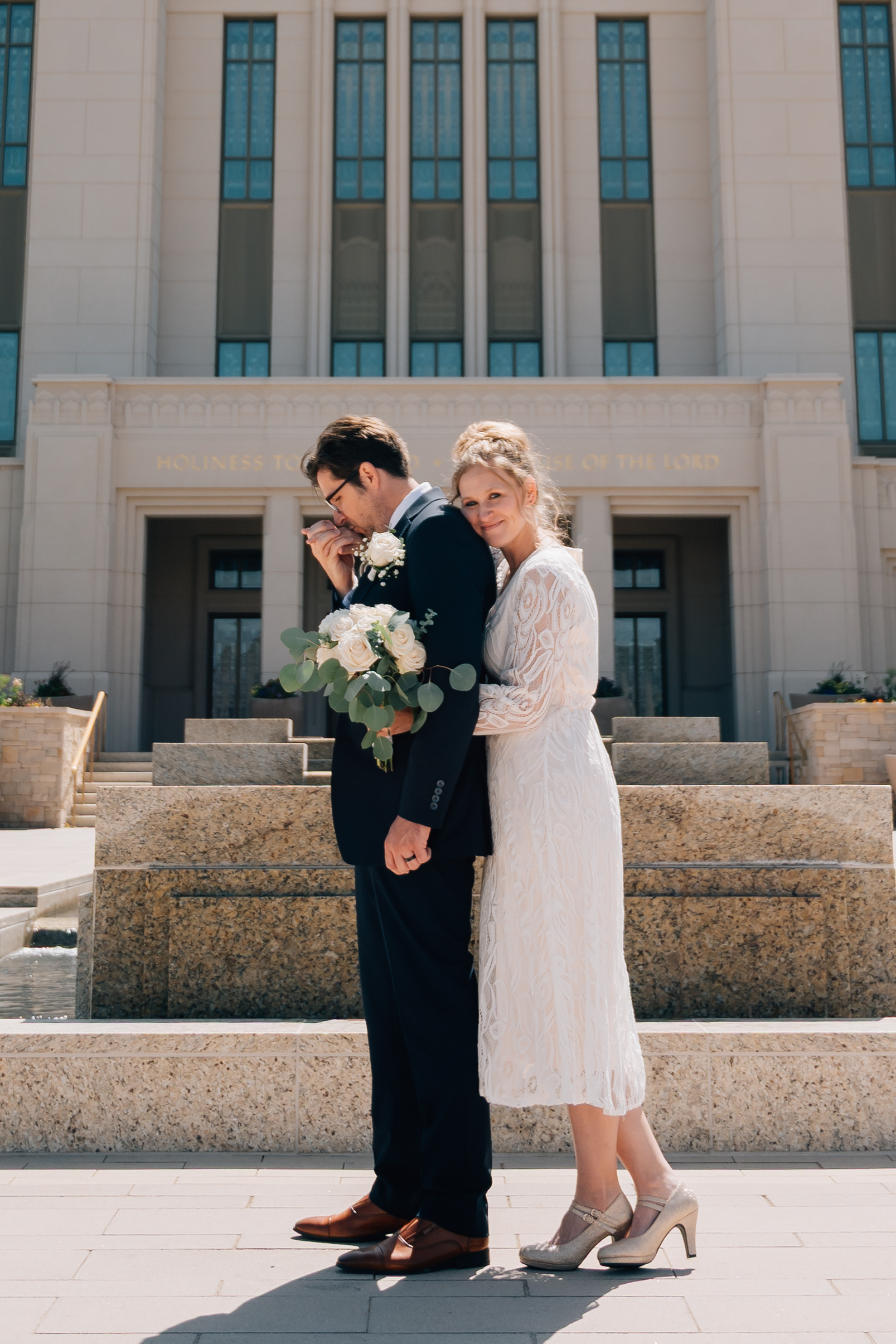 Groom kisses his brides hand in front of the LDS temple in Saratoga Springs Ut while she cuddles up from behind him.