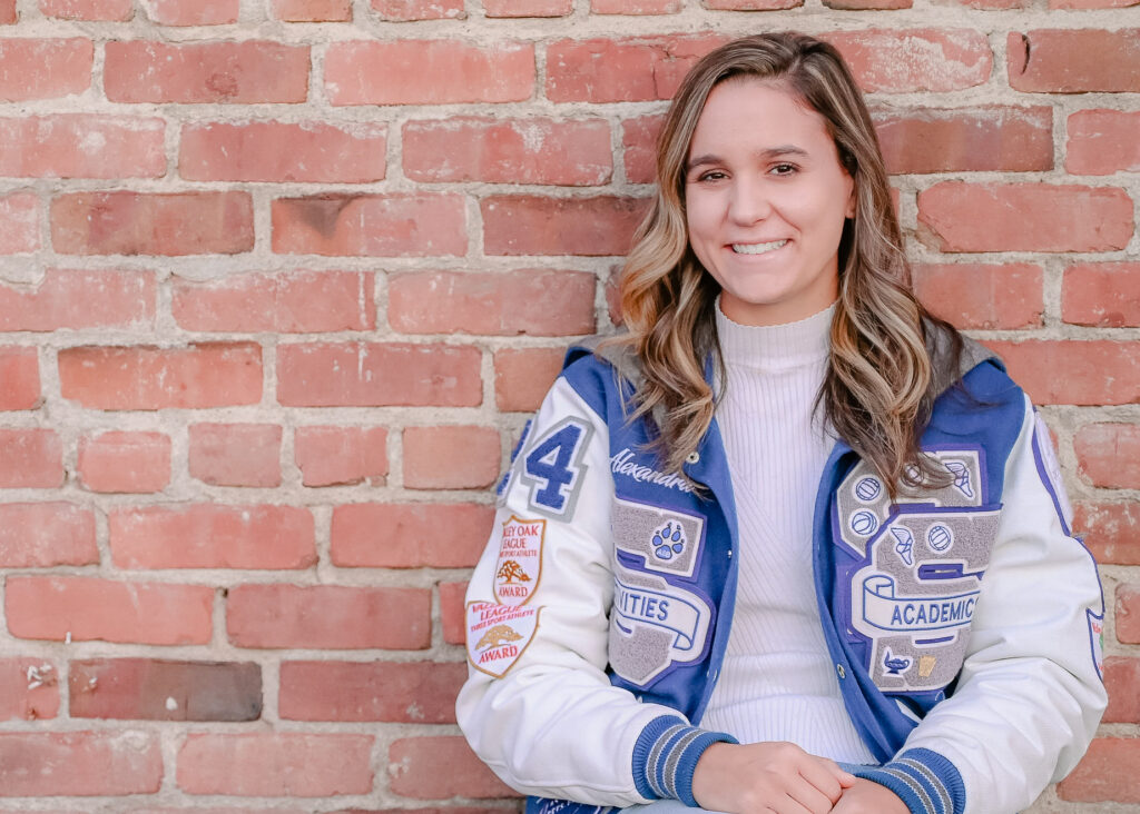 A senior girl wearing her letterman jacket while sitting with her hands in her lap in front of a brick wall while she smiles on.