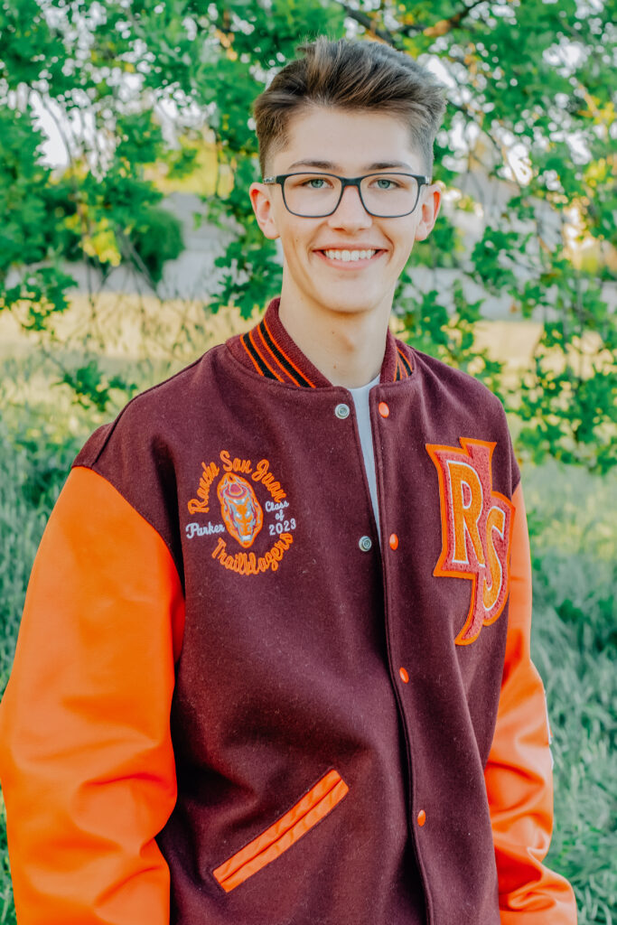 A graduating senior boy smiles while wearing his letterman jacket in front of a tree.
