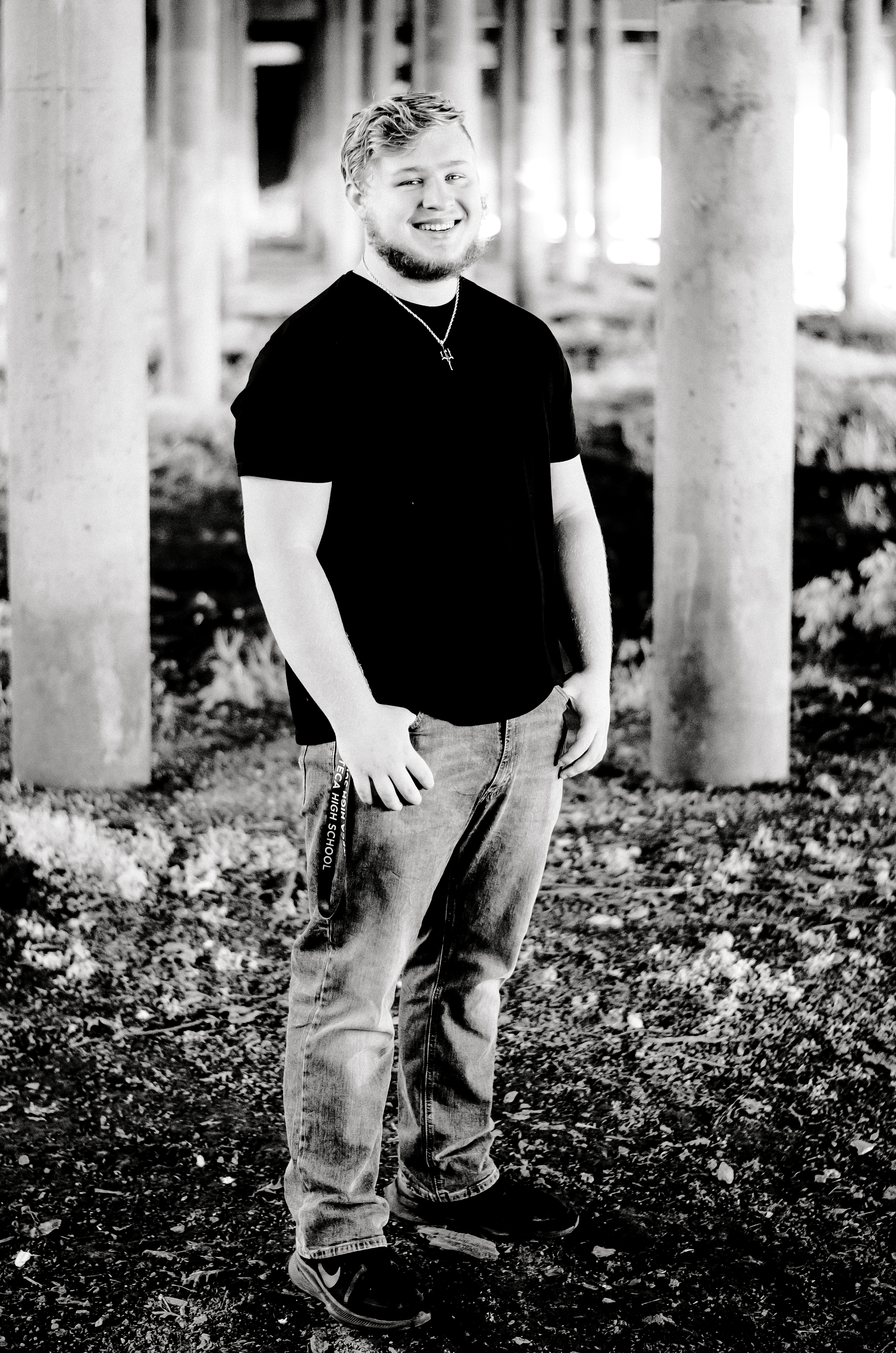 A boy with thumbs in pockets smiles while posing to the side under the bridge. 