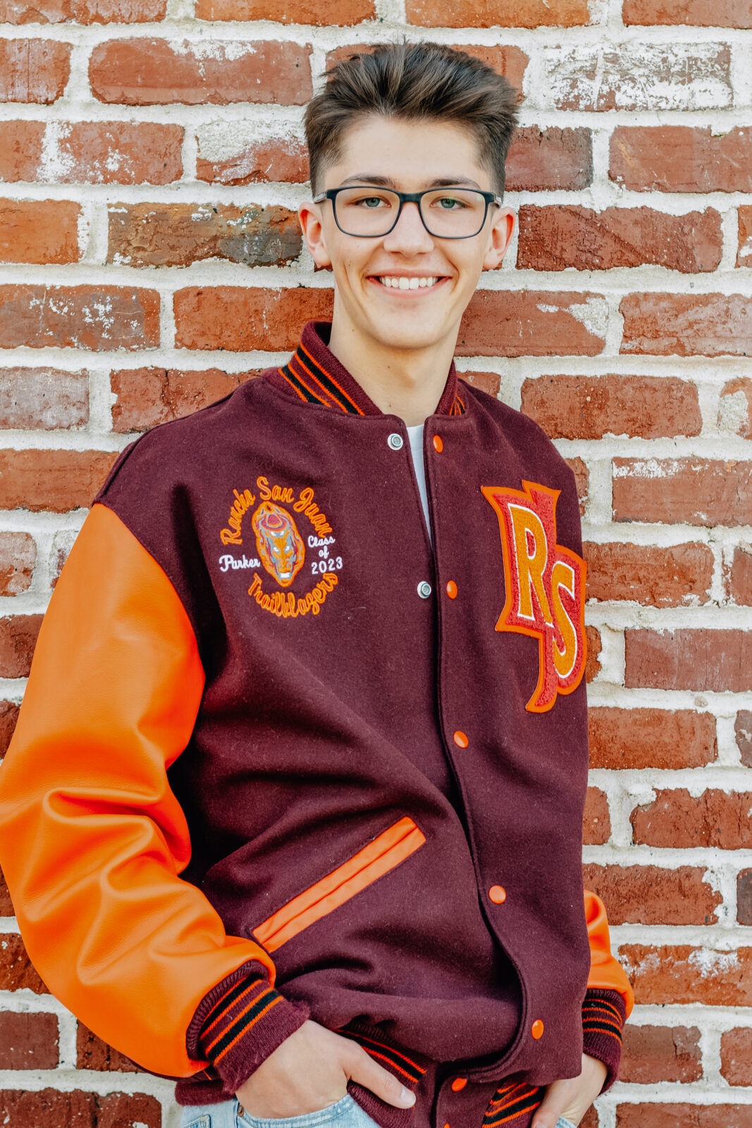 A graduating senior boy smiles while wearing his letterman jacket with his hands in his pockets in front of a brick wall in Ripon California.