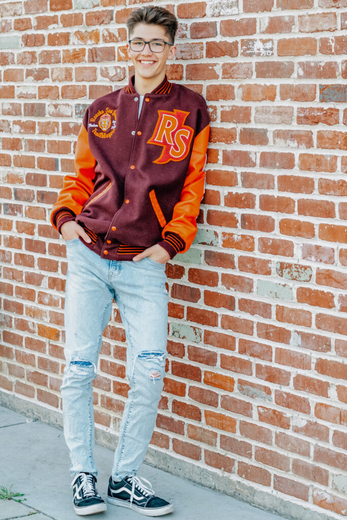 A graduating senior boy smiles while wearing his letterman jacket with his hands in his pockets in front of a brick wall in Ripon California.