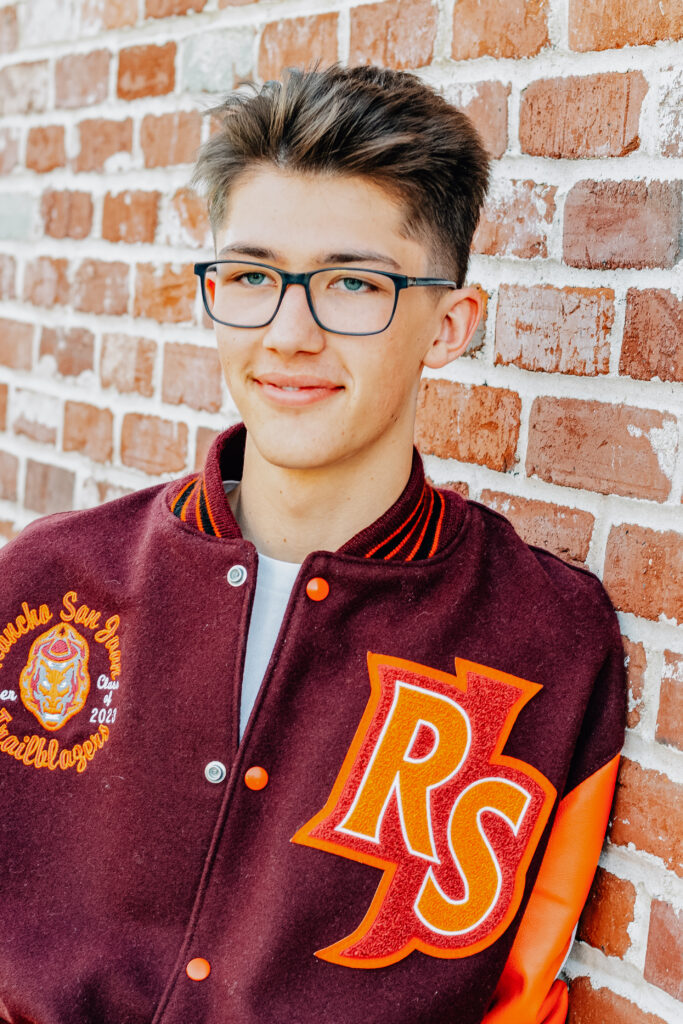 A graduating senior boy smiles while wearing his letterman jacket with one hand in a pocket and looking off to the side. The golden hour hitting the brick wall in the background at the Ripon, CA castle.
