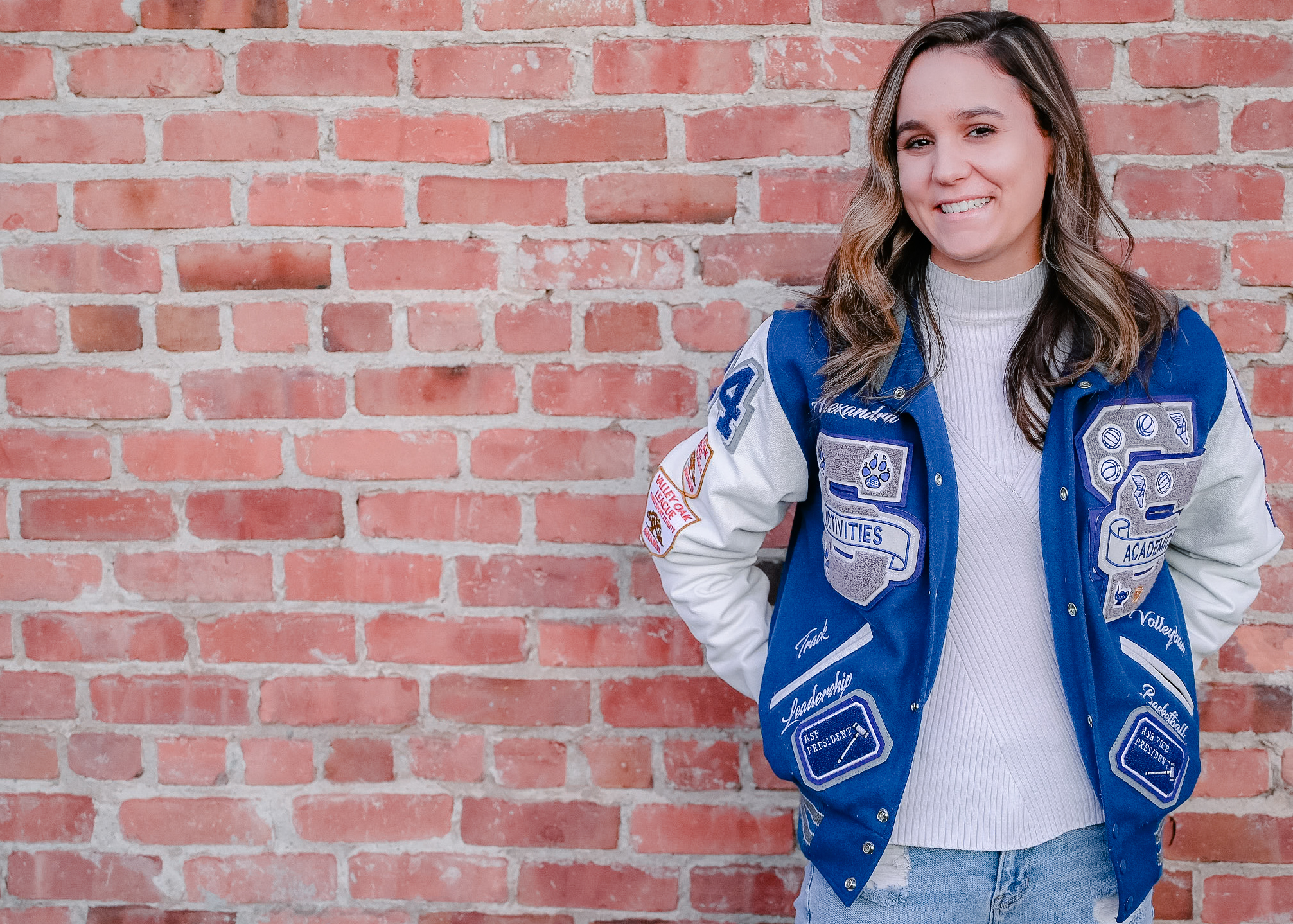A senior girl wearing her letterman jacket with her hands in her back pockets while posing in front of a brick wall while she smiles on.