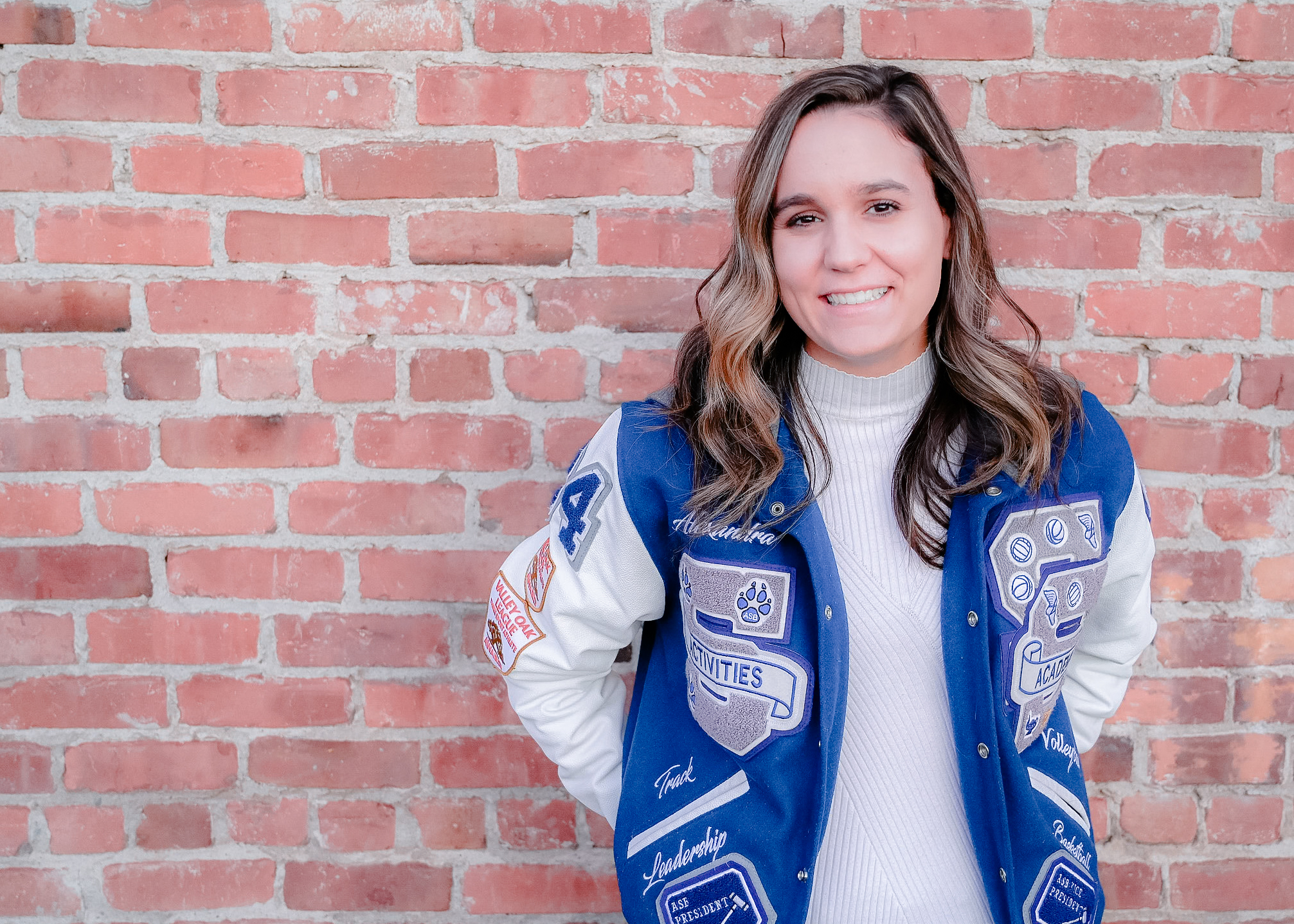 A senior girl wearing her letterman jacket with her hands in her back pockets while she smiles on.