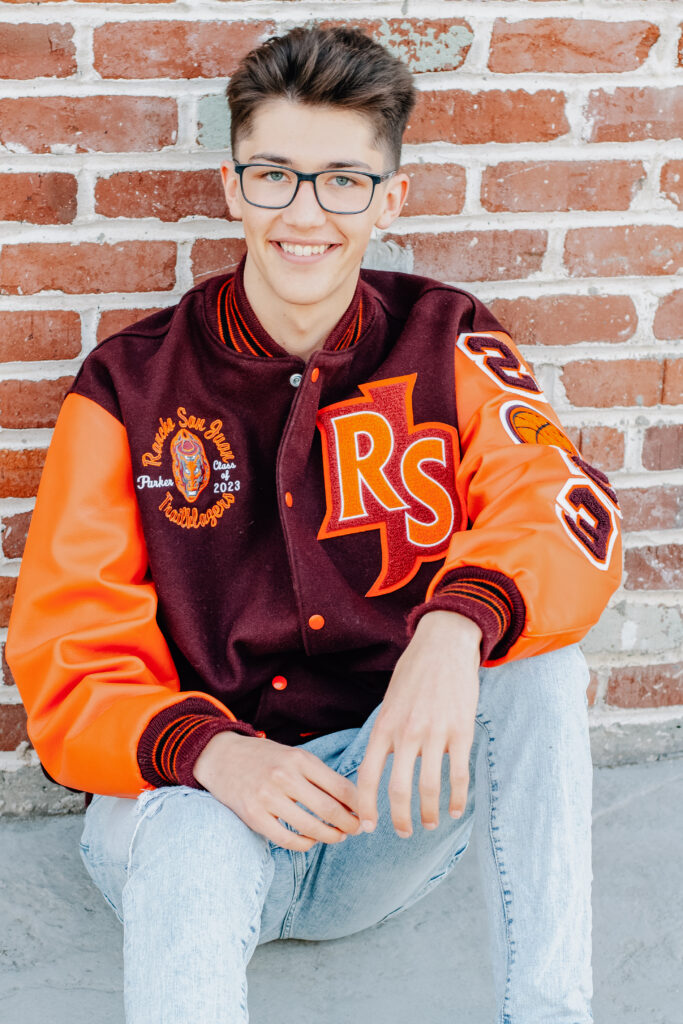 A senior boy sitting in front of a brick wall wearing his letterman jacket with his knees bent and his hands resting in between them.