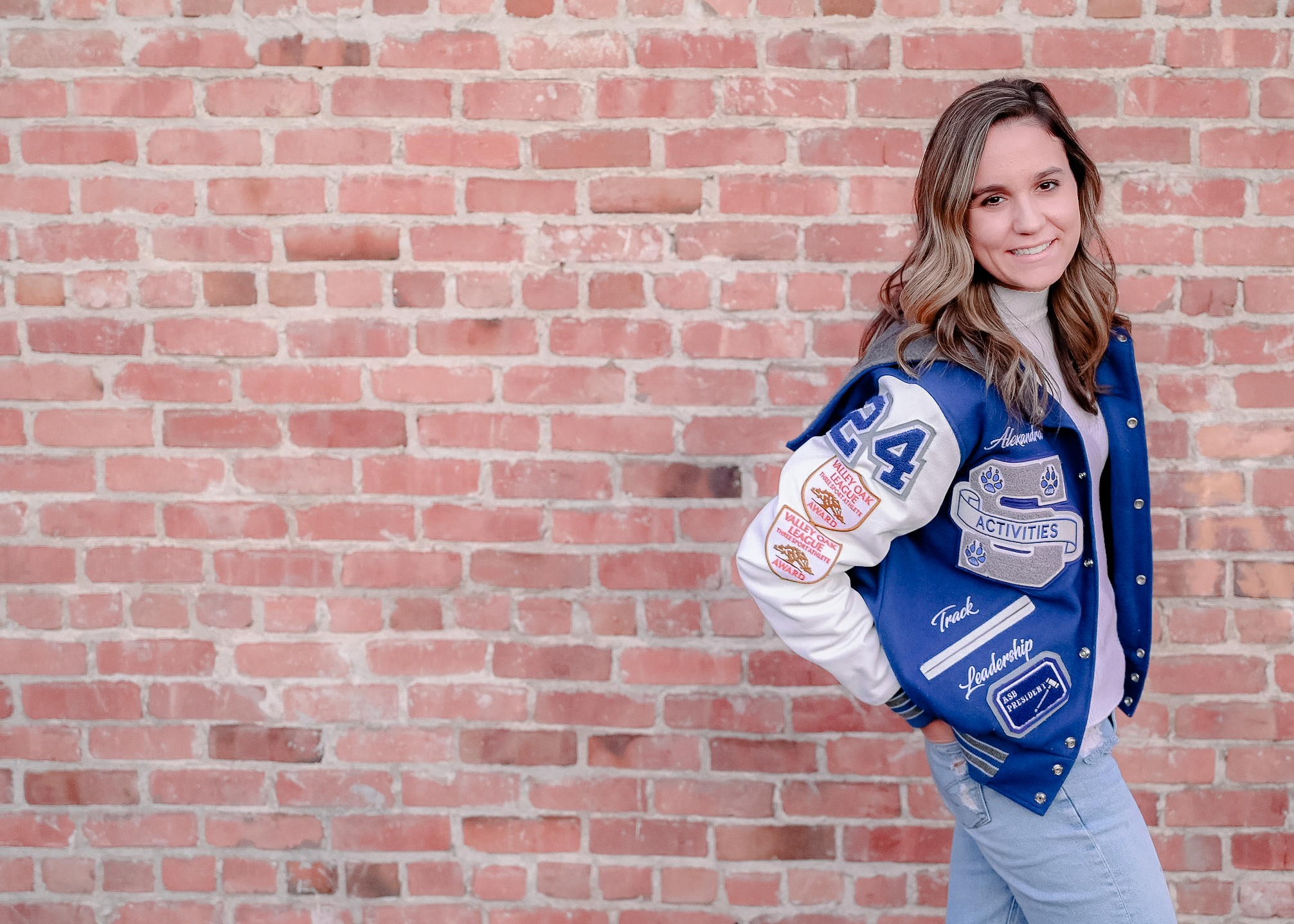 A senior girl wearing her letterman jacket with her hands in her back pockets while posing in front of a brick wall while she smiles on.