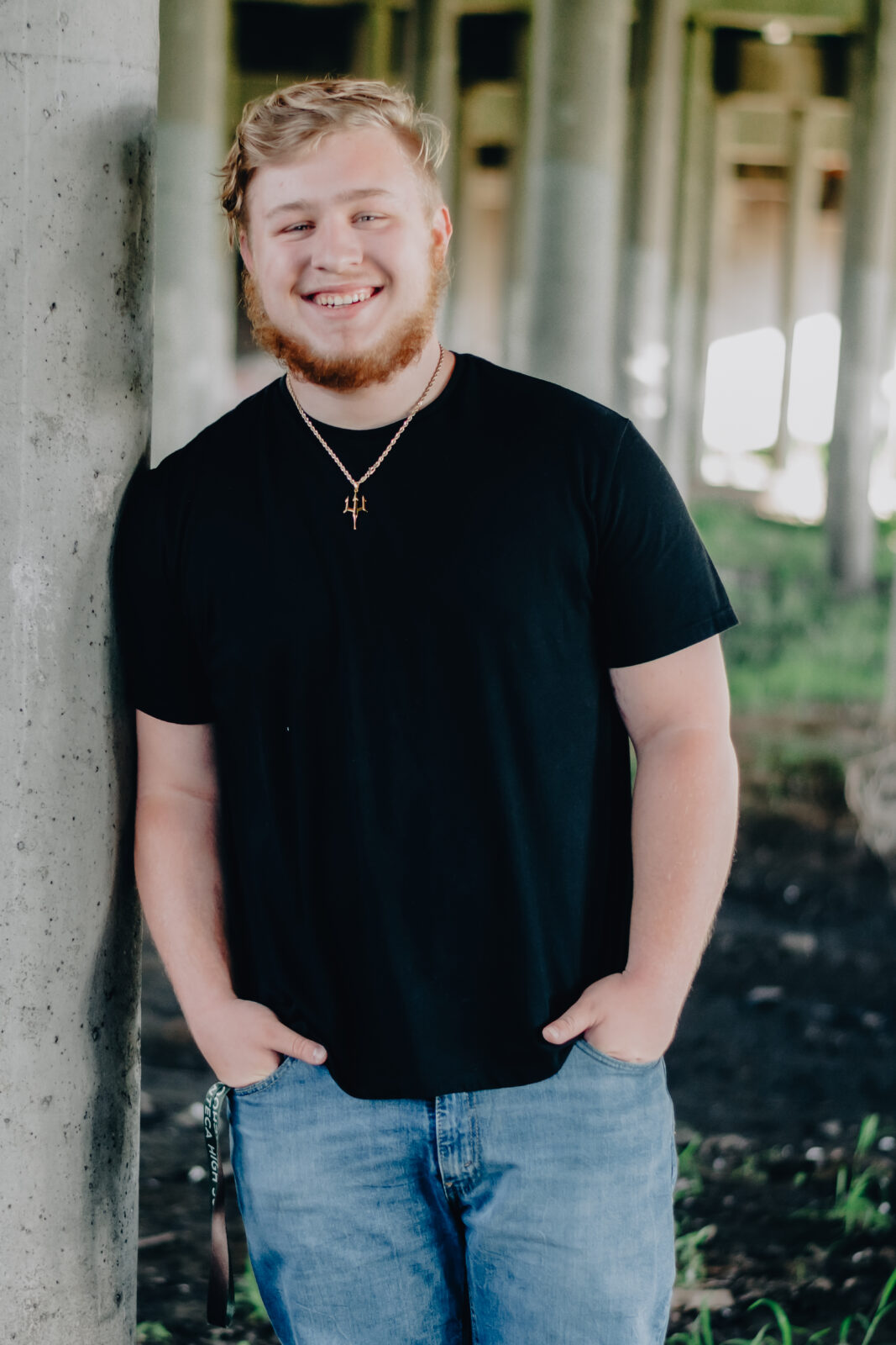 A graduating senior boy smiles under the bridge while leaning up against a cement post Hands in pockets and a big smile on his face.