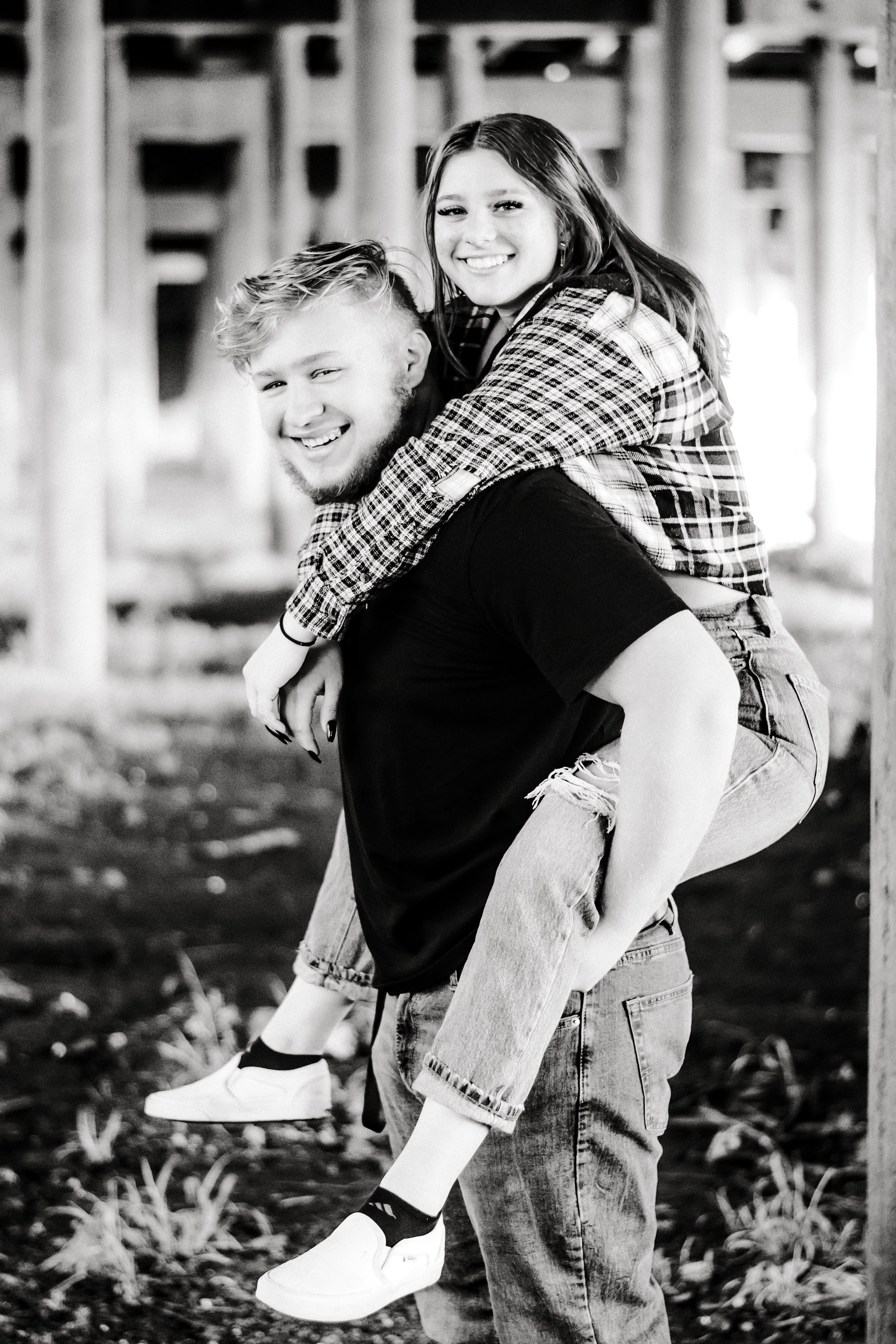 A Senior graduating boy has his girlfriend on his back. She has her arms around his neck while looking relaxed and smiling on. 