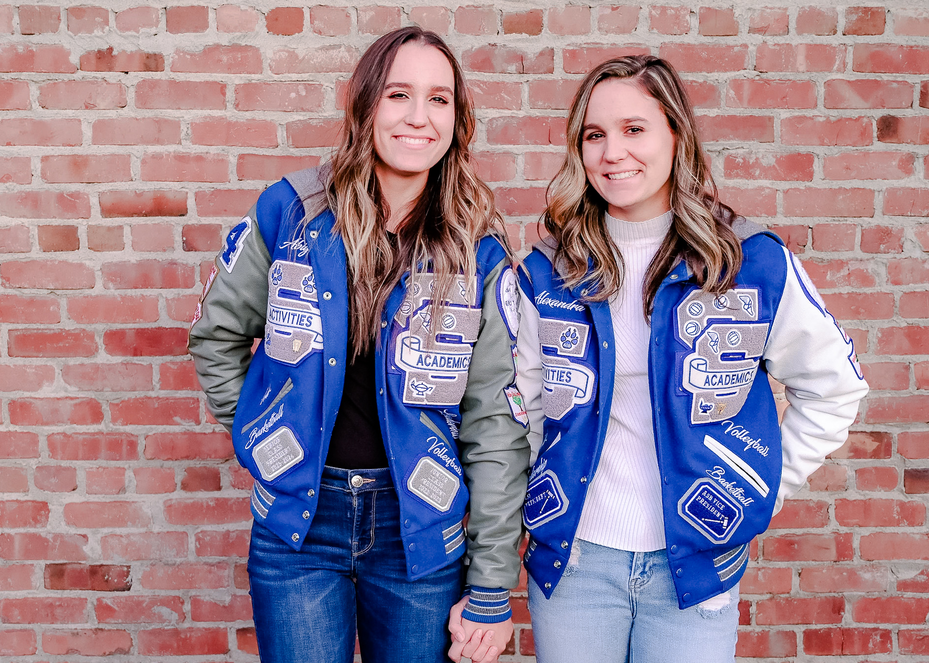 Twin Senior girls holding hands while posing in their letterman jackets in front of a brick wall.