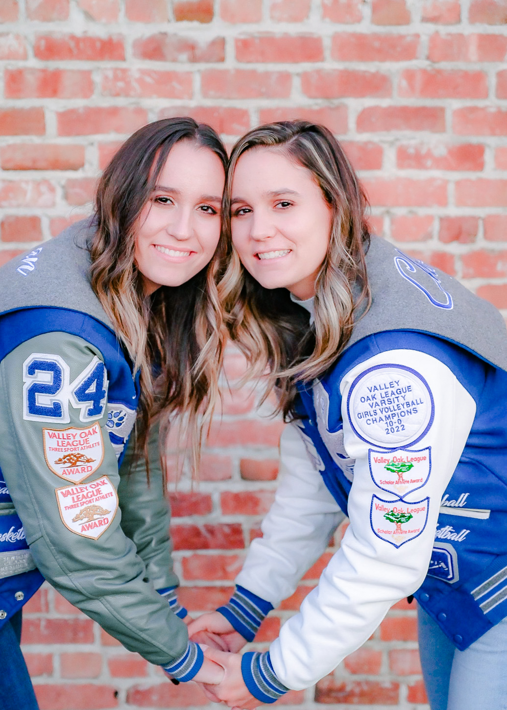 Twin Senior girls holding hands while posing in their letterman jackets in front of a brick wall.