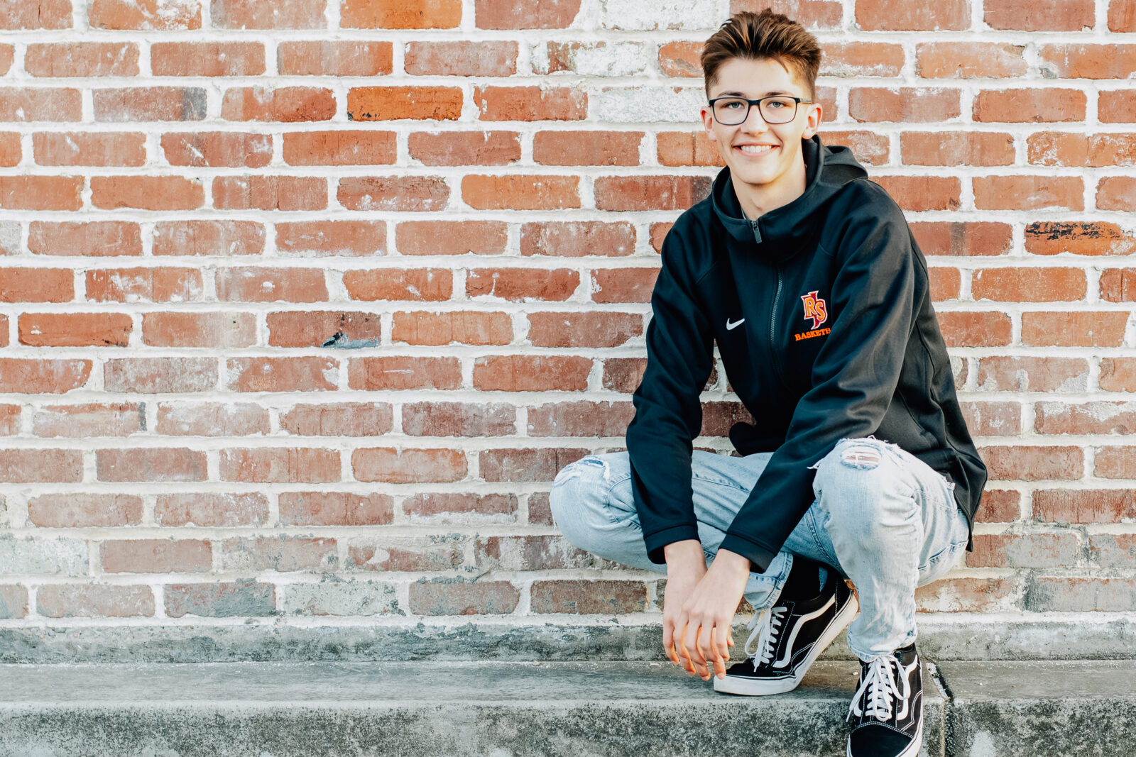 A senior boy crouching down in front of a brick wall wearing his Rancho San Juan High School basketball hoodie with his knees bent and his hands resting in between them. Feeling relaxed as the sun goes down on Ripon, CA.
