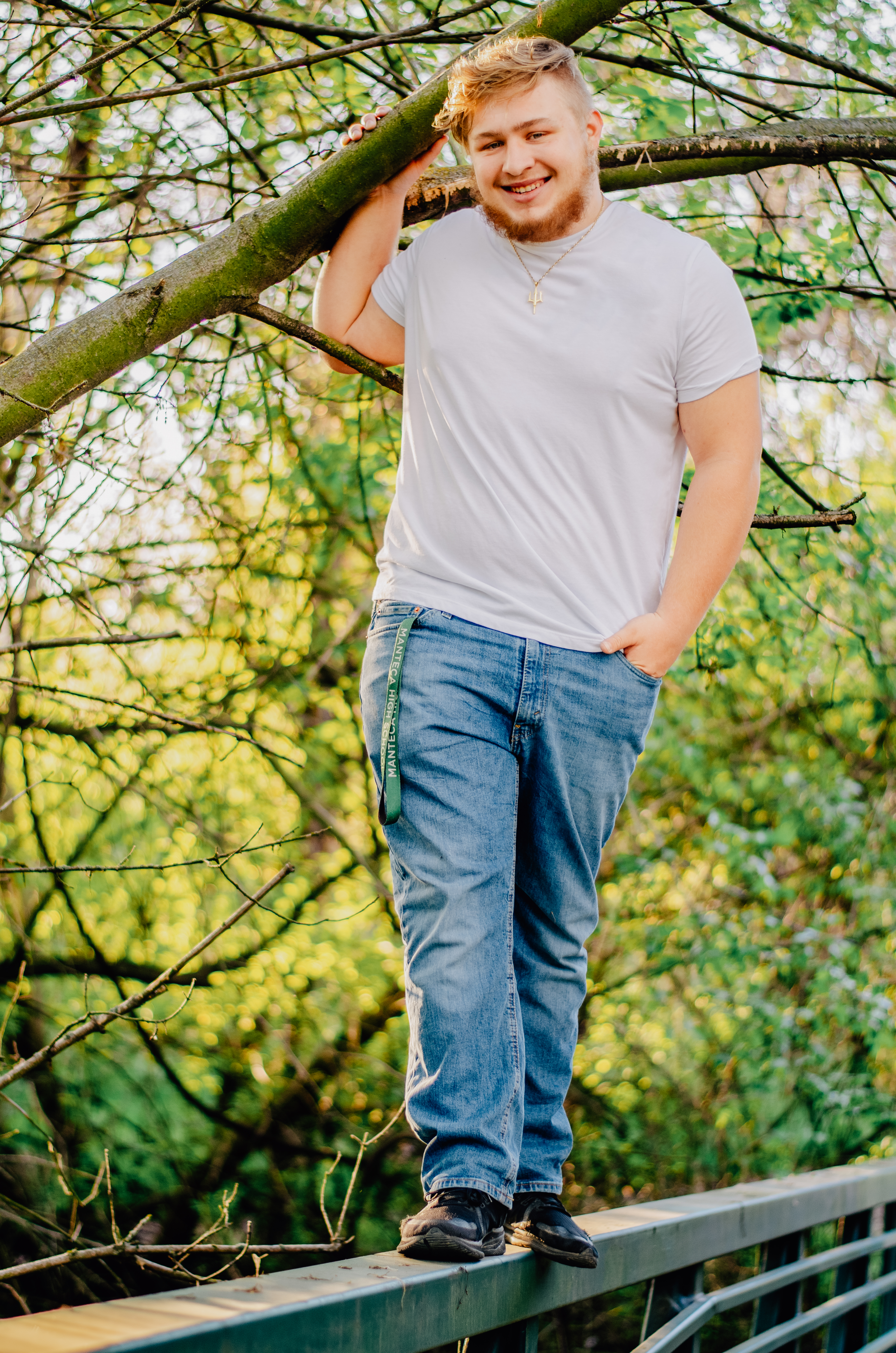 Senior boy hops up on the bridge and stands with success while holding onto a tree branch with one hand in his pocket and the other safely on a tree branch to keep him steady.  