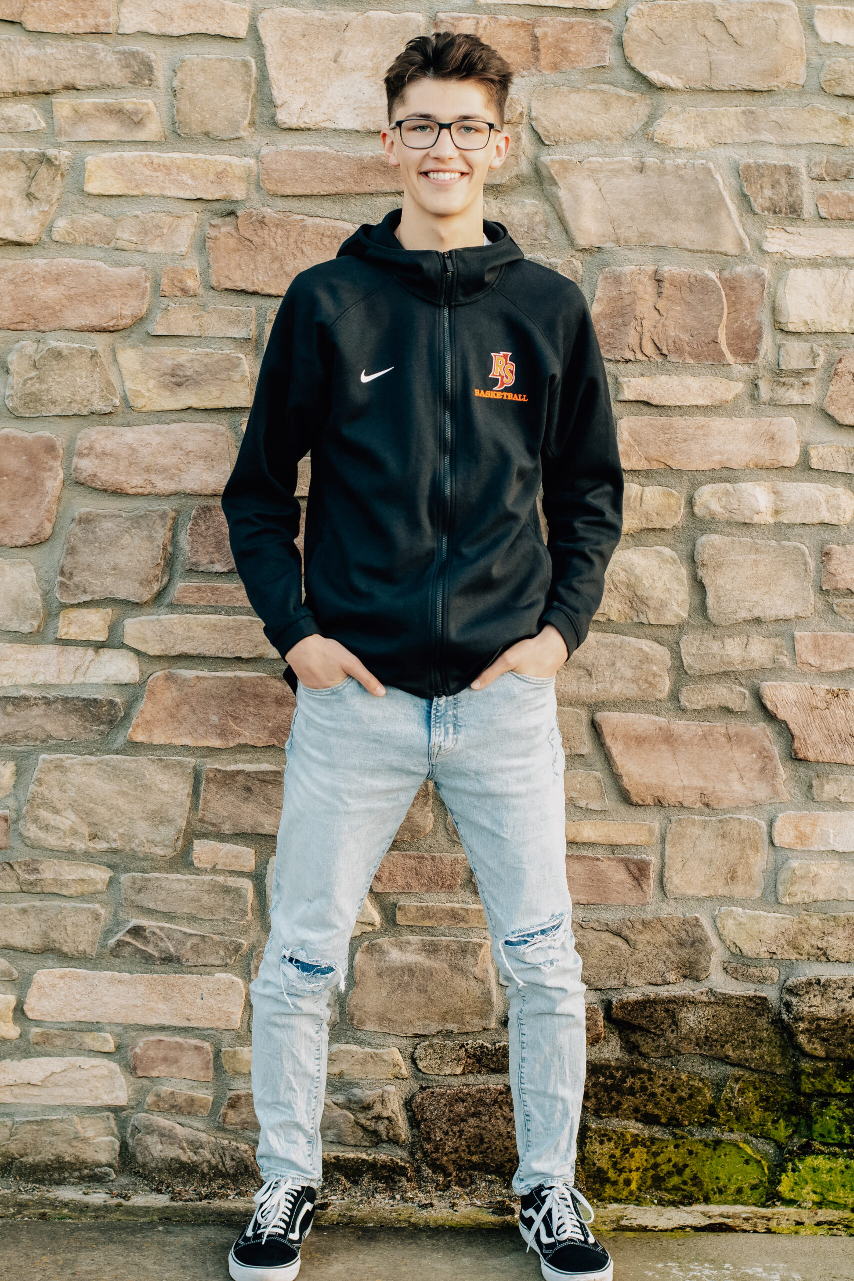 A senior boy graduating from Rancho San Juan High School wears his basketball warm up sweater shirt with his hands in his pockets and smiling.