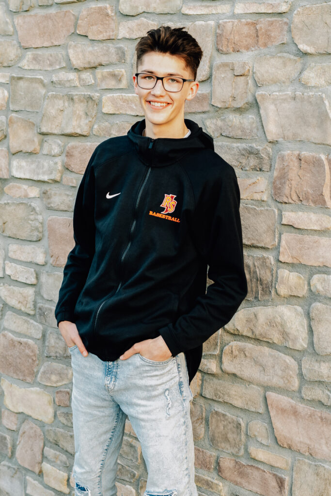 A Senior boy smiles with his hands in his pockets while in front of a stone wall at the Ripon, CA castle downtown.