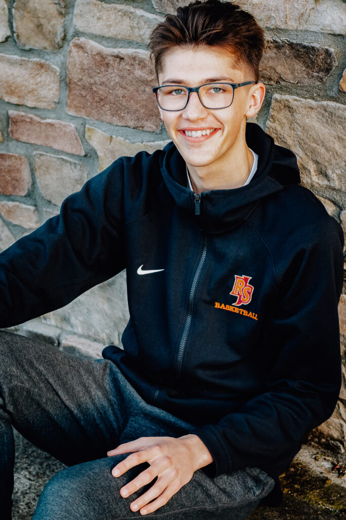 A senior boy sits in front of a stone wall with his hands relaxed on his legs at the Ripon Castle in California.