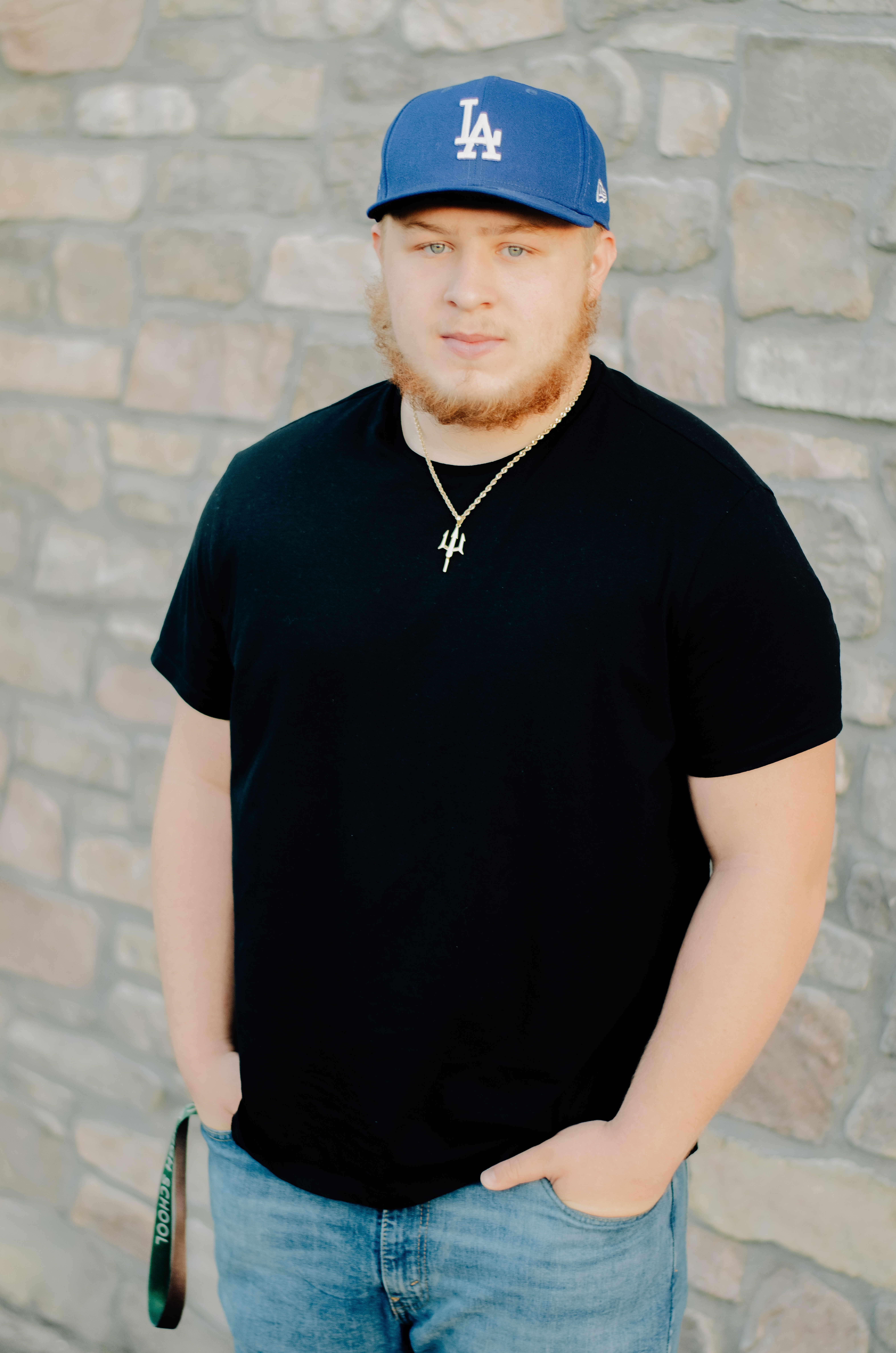 A boy leaned by a stone wall with hands in his front pockets and wearing a LA Dodgers ball cap giving off a smoulder vibe.