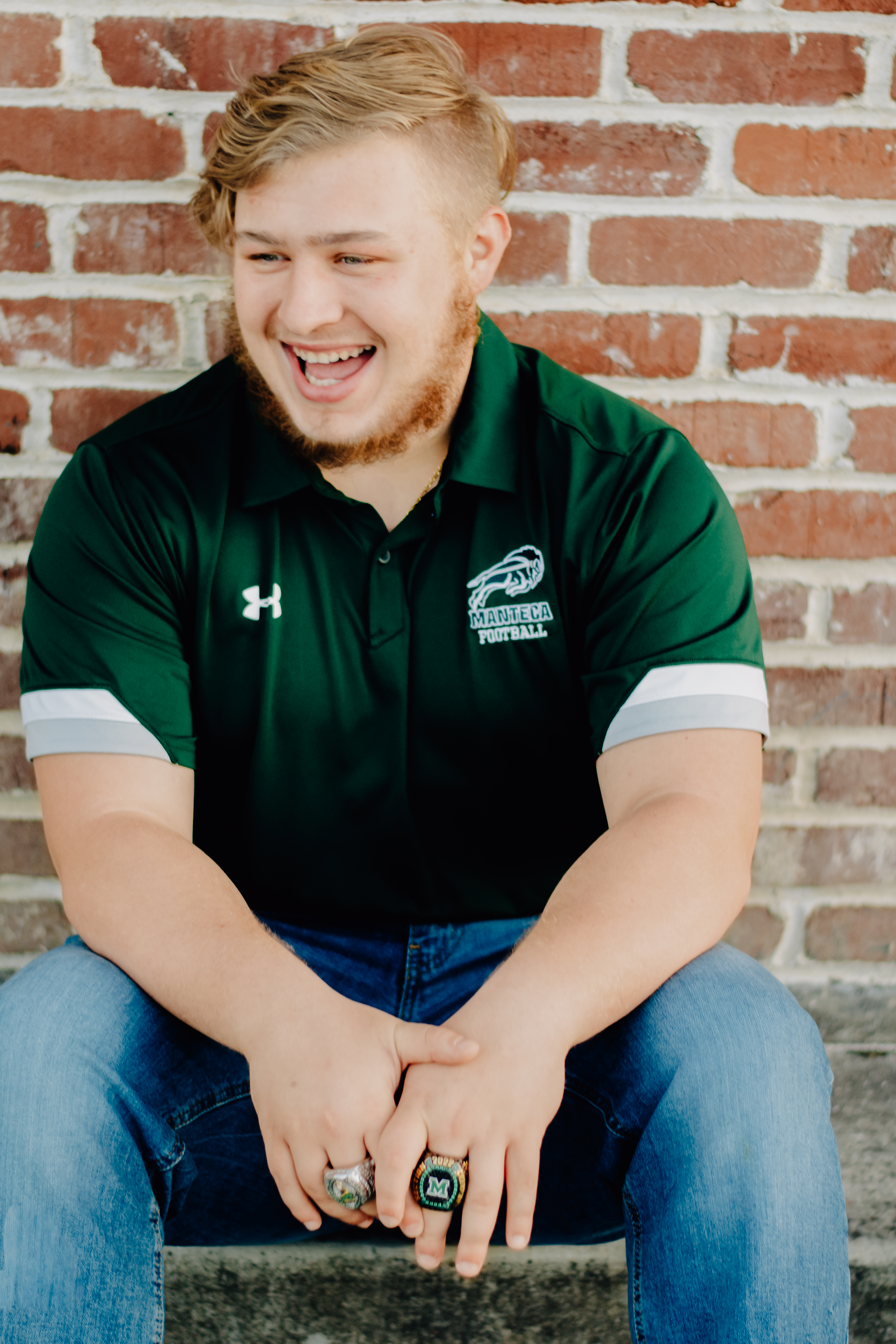 A boy sitting in front of a brick wall and leaning forward with hands relaxed in front of him. Showing off his Championship rings from Manteca High School Football