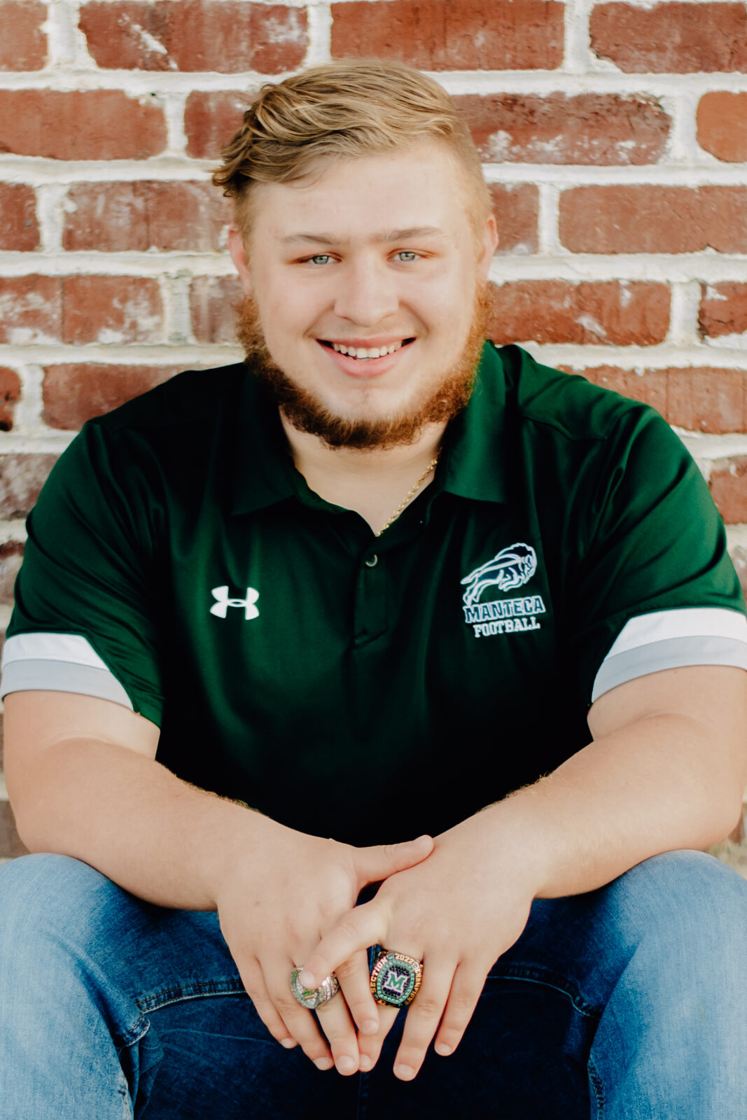 A boy sitting in front of a brick wall and leaning forward with hands relaxed in front of him. Showing off his Manteca Buffs Football Championship rings.