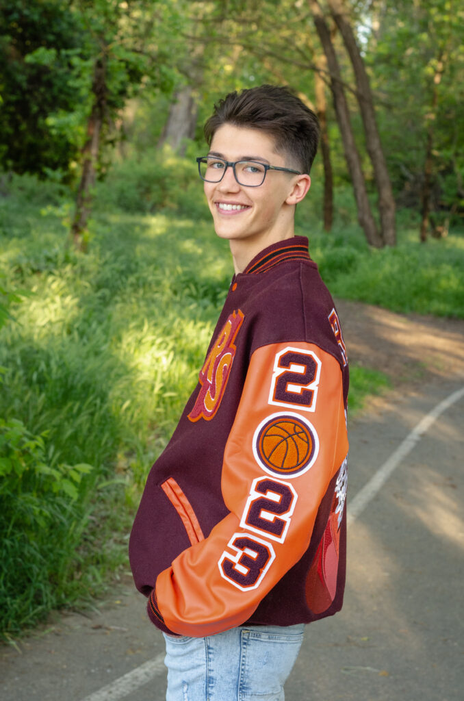 A graduating senior boy smiles while wearing his letterman jacket in front of a tree.