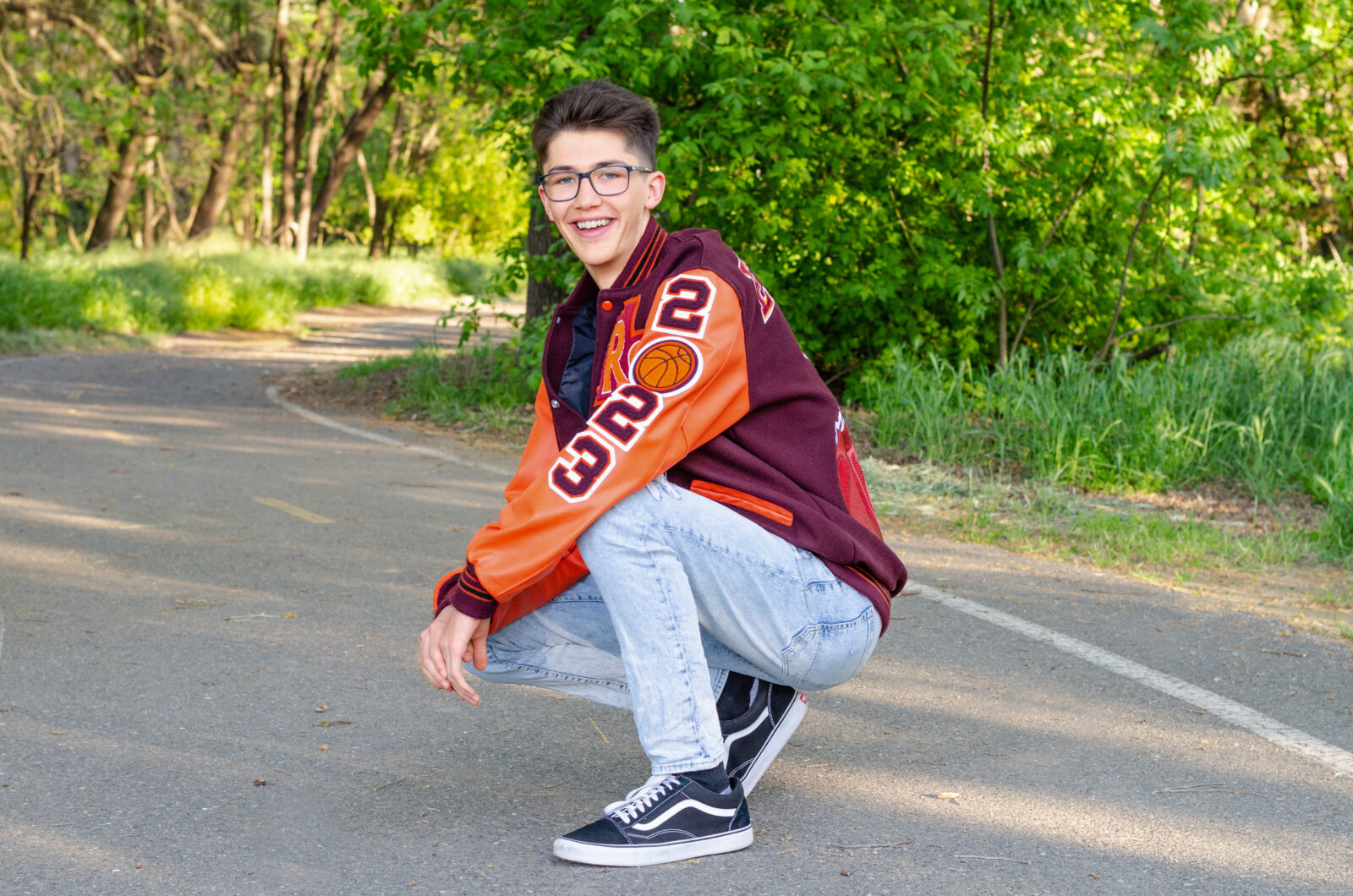 A graduating senior boy smiles while wearing his letterman jacket on a path. He is crouched down with his relaxed in between his knees.