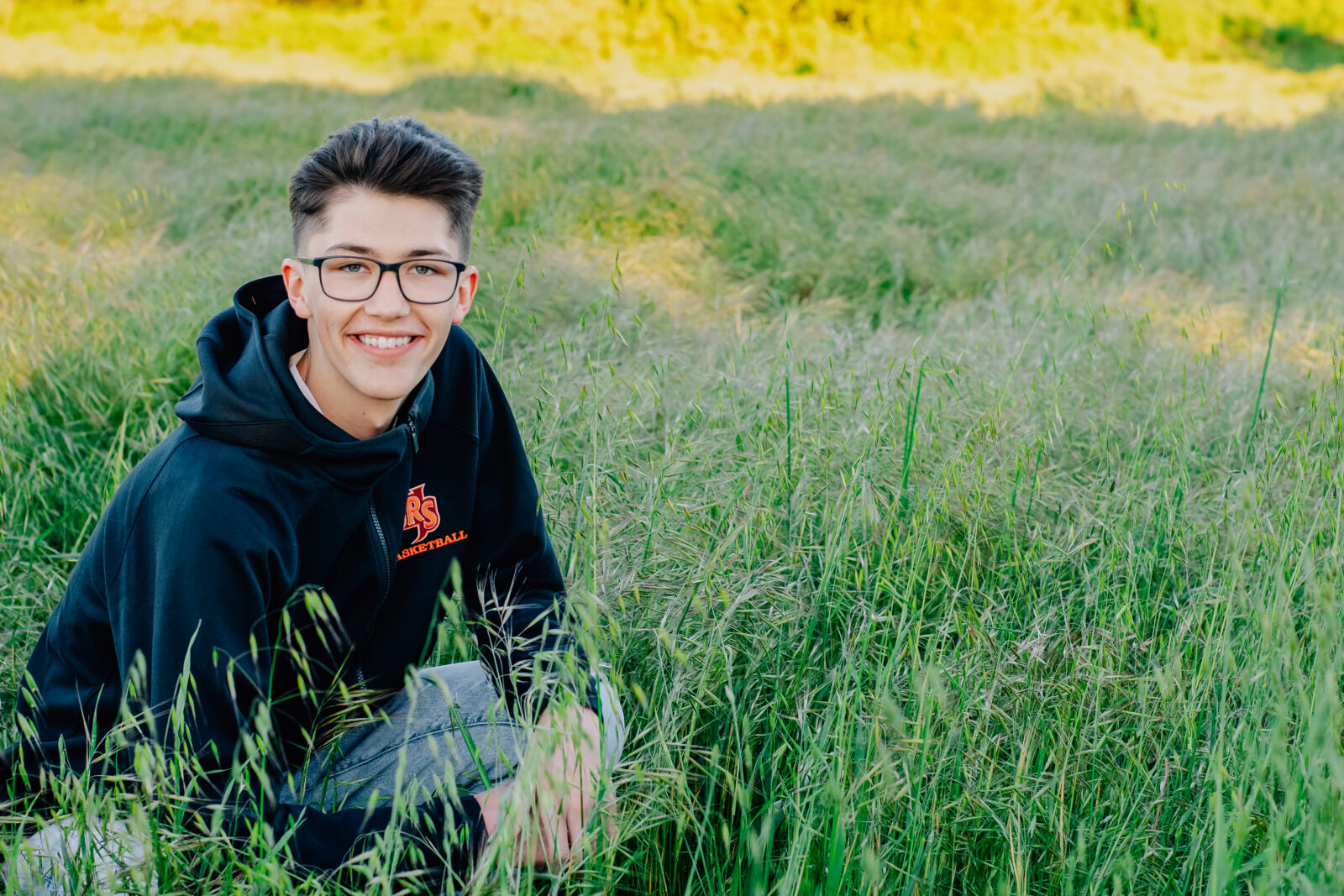 A graduating senior boy smiles while wearing his black basketball hoodie while crouched down in a field. Feeling relaxed at this golden hour session.