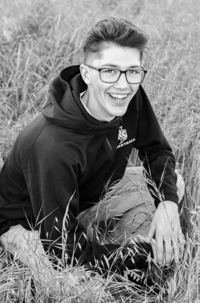 A senior boy crouching down in a field wearing his Rancho San Juan High School basketball hoodie with his knees bent and his hands resting in between them. Feeling relaxed as the sun goes down on Army Corps Park in Ripon, CA.