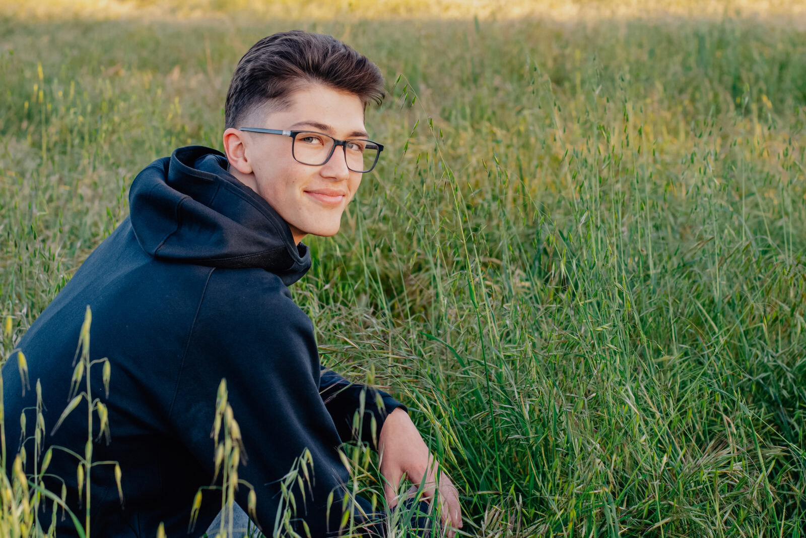 A graduating senior boy smiles while wearing his black basketball hoodie while crouched down in a field. Feeling relaxed at this golden hour session.