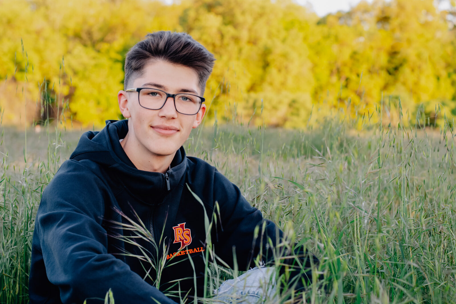A graduating senior boy smiles while wearing his black basketball hoodie while sitting down in a field. Feeling relaxed at this golden hour session.