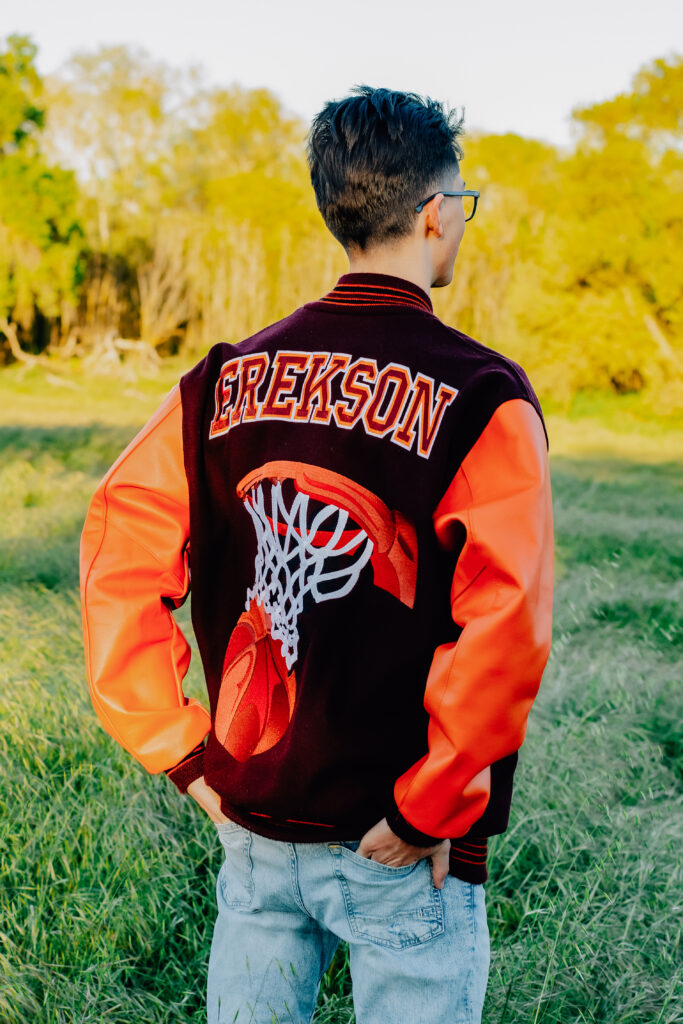 A graduating senior boy shows off his last name on the back of his letterman jacket in front of an open field at sunset.