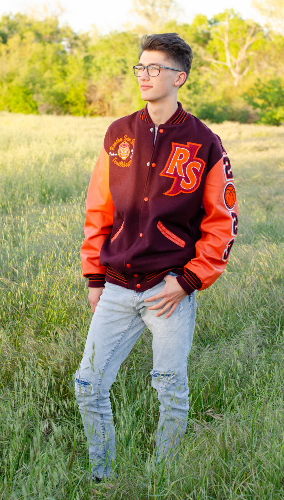 A graduating senior boy smiles while wearing his letterman jacket with one hand in a pocket and looking off to the side. The golden hour hitting the field and trees in the background.