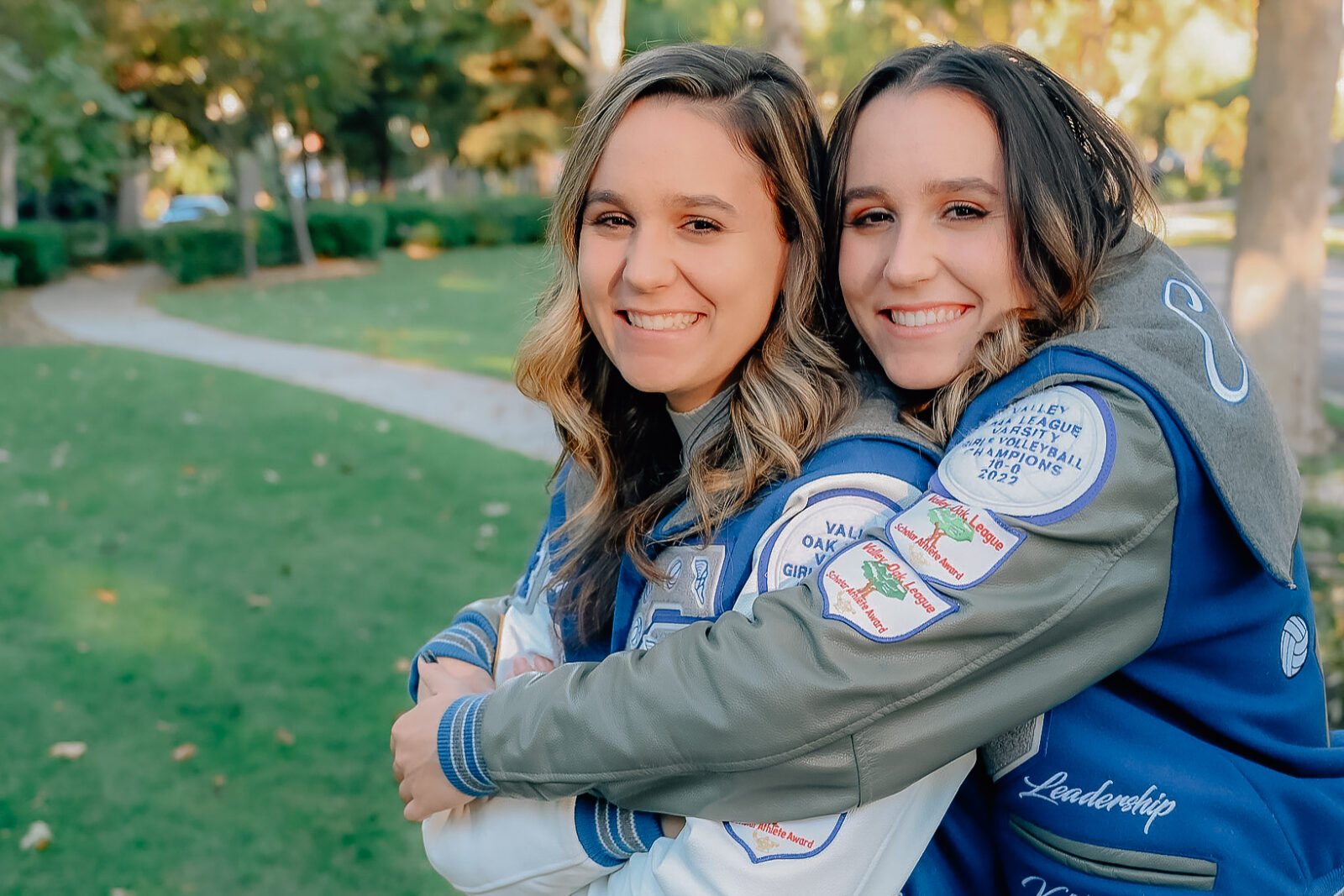 Twin girls in letterman jackets. One twin is embracing the other from behind the back while the twin in the front has her arms crossed. Both girls laughing for the camera.