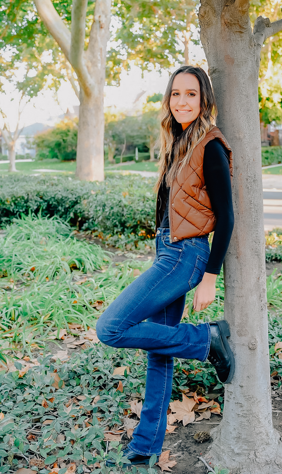 A senior girl resting against a tree trunk while smiling for the camera with her hand rests next to her leg that is up against the tree.