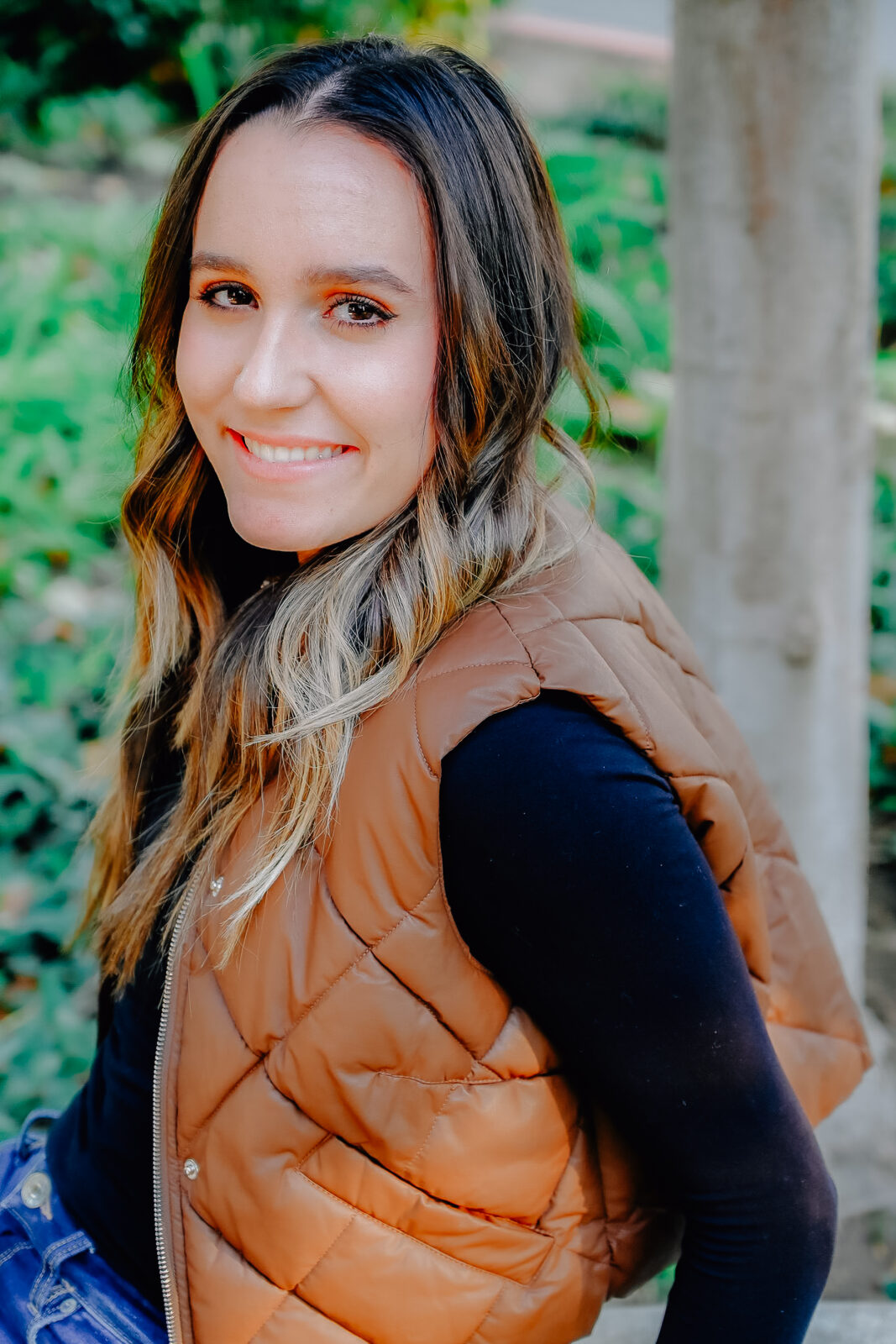 A senior girl wearing a long black shirt with a brown vest. Her hands behind her resting on a bench while she smiles on for a close up.