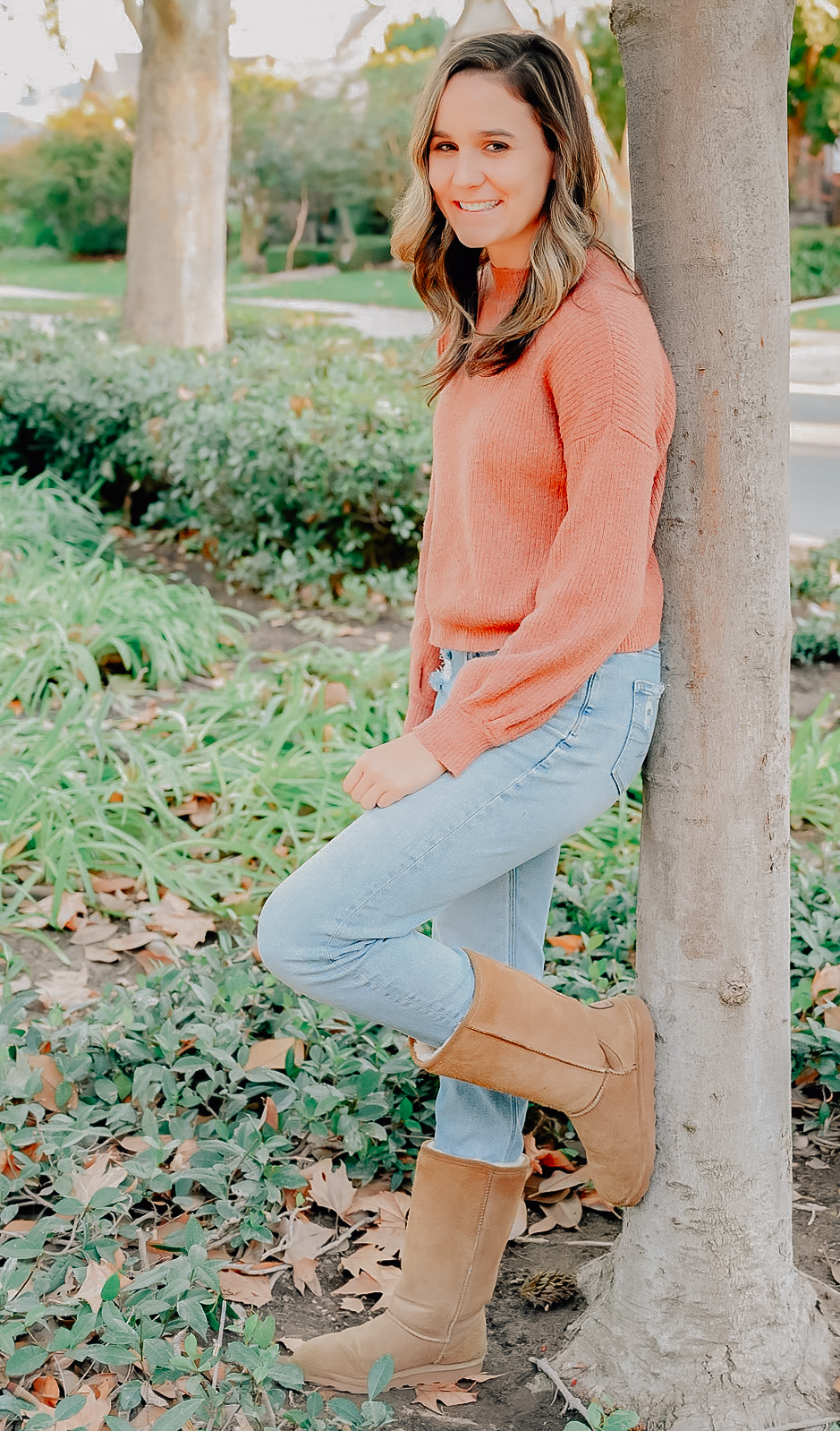 A senior girl resting against a tree trunk while smiling for the camera with her hand on her bent leg that is up against the tree.