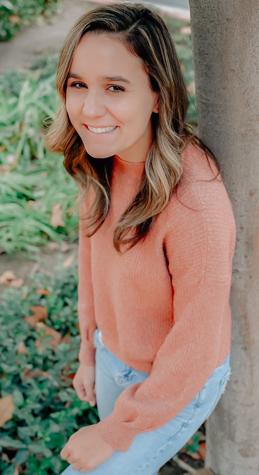A senior girl resting against a tree trunk while smiling for the camera with her hand on her bent leg that is up against the tree.