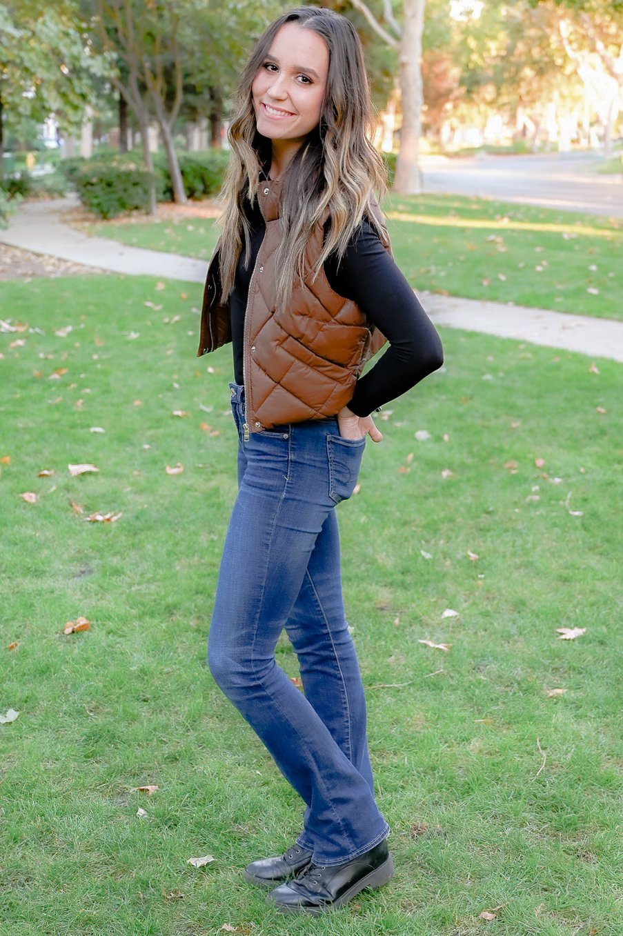 A senior girl wearing a long black shirt with a brown vest. Her hands in her back pockets posing in a neighborhood while she smiles on.