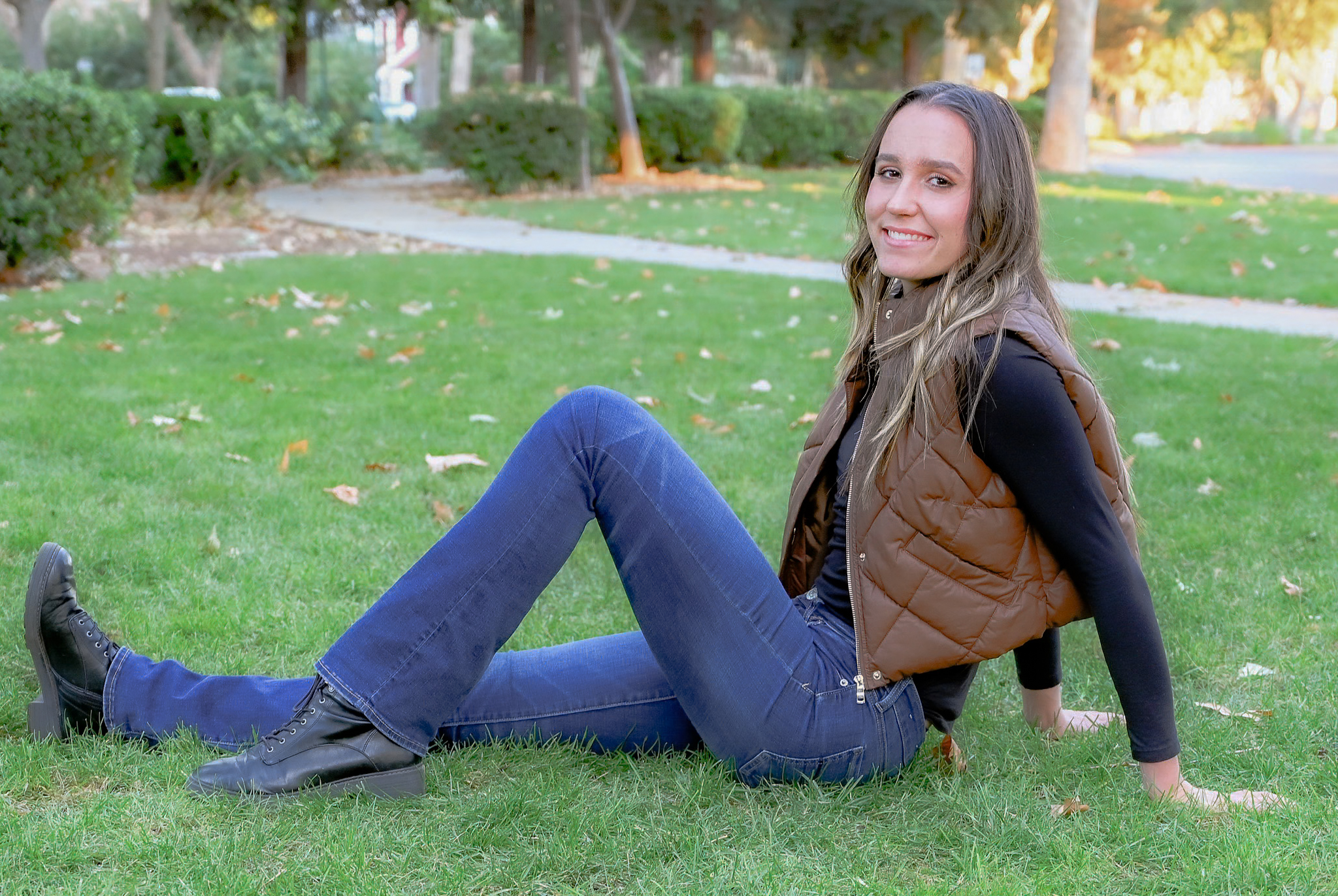 A senior girl posing on grass with one leg extended and one leg angled resting her hands behind her on the grass while she smiles for the camera.