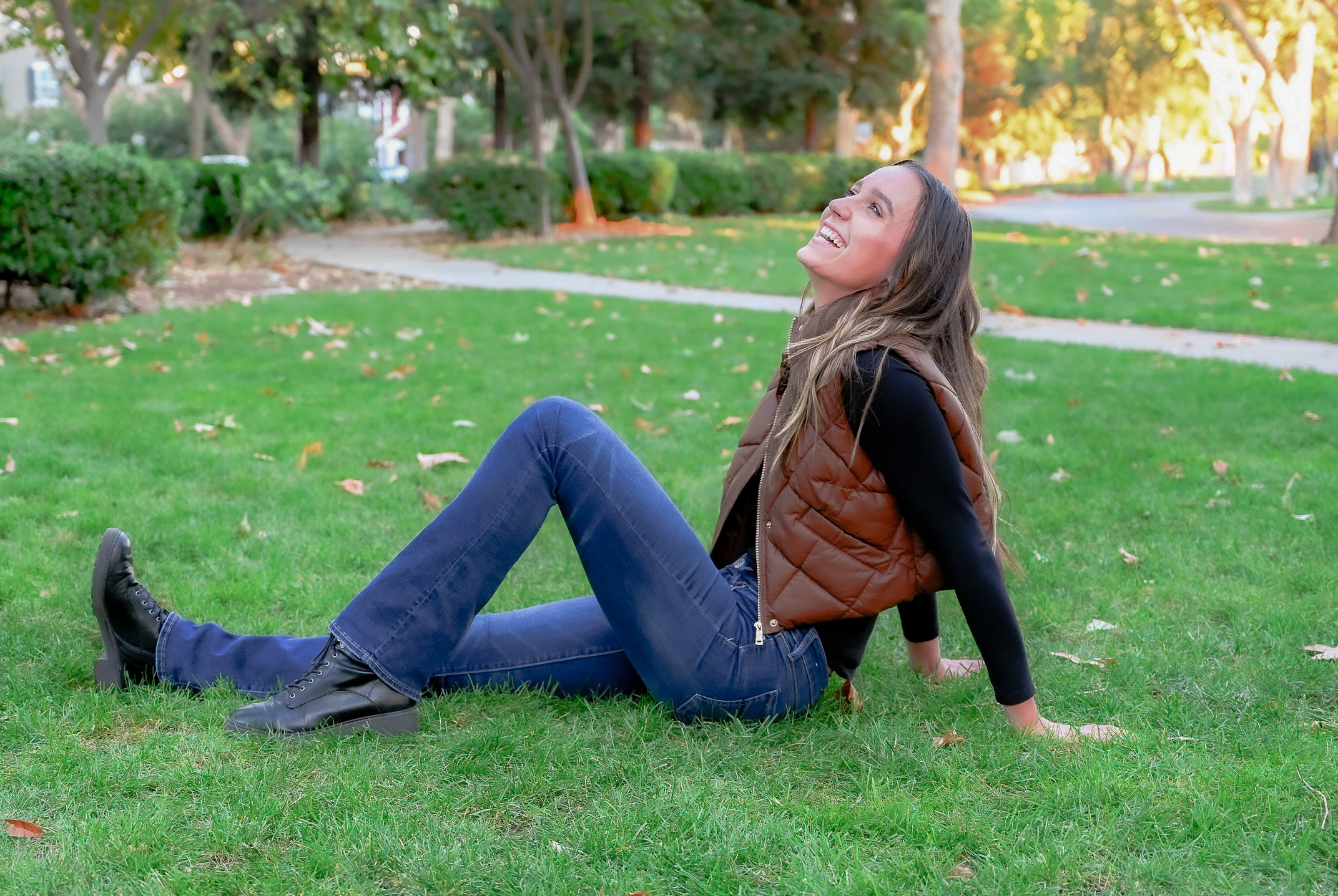 A senior girl posing on grass with one leg extended and one leg angled resting her hands behind her on the grass while she laughs for the camera.