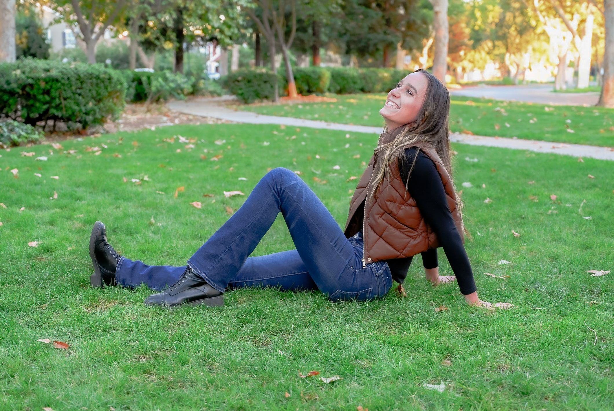 A senior girl posing on grass with one leg extended and one leg angled resting her hands behind her on the grass while she laughs for the camera.