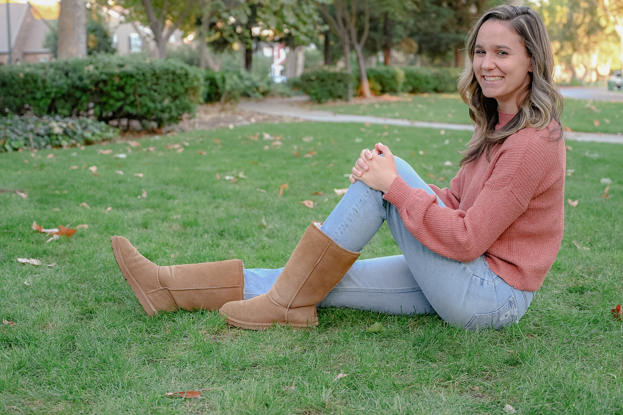 A senior girl posing on grass with one leg extended and one leg angled resting her hands on her knee while she laughs for the camera.