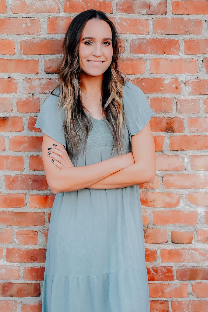 A senior girl wearing a mint colored dress while leaning up against a brick wall folding her arms in front of her and smiling towards the camera.