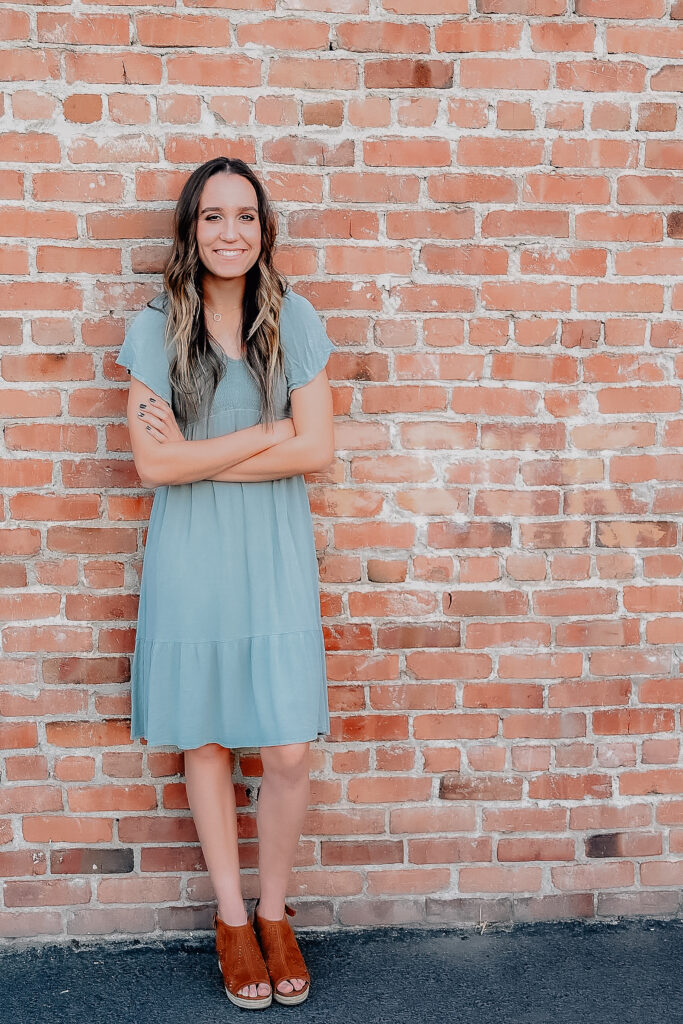 A senior girl wearing a mint colored dress while leaning up against a brick wall folding her arms in front of her and smiling towards the camera.
