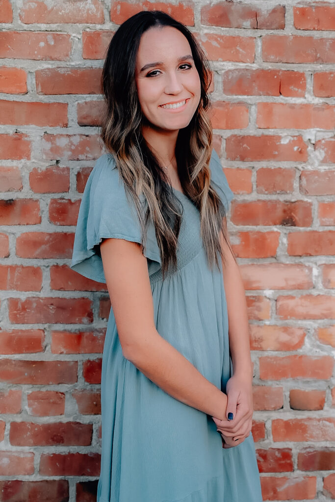 A senior girl wearing a mint colored dress while leaning up against a brick wall hands crossed at her waist and feeling relaxed.