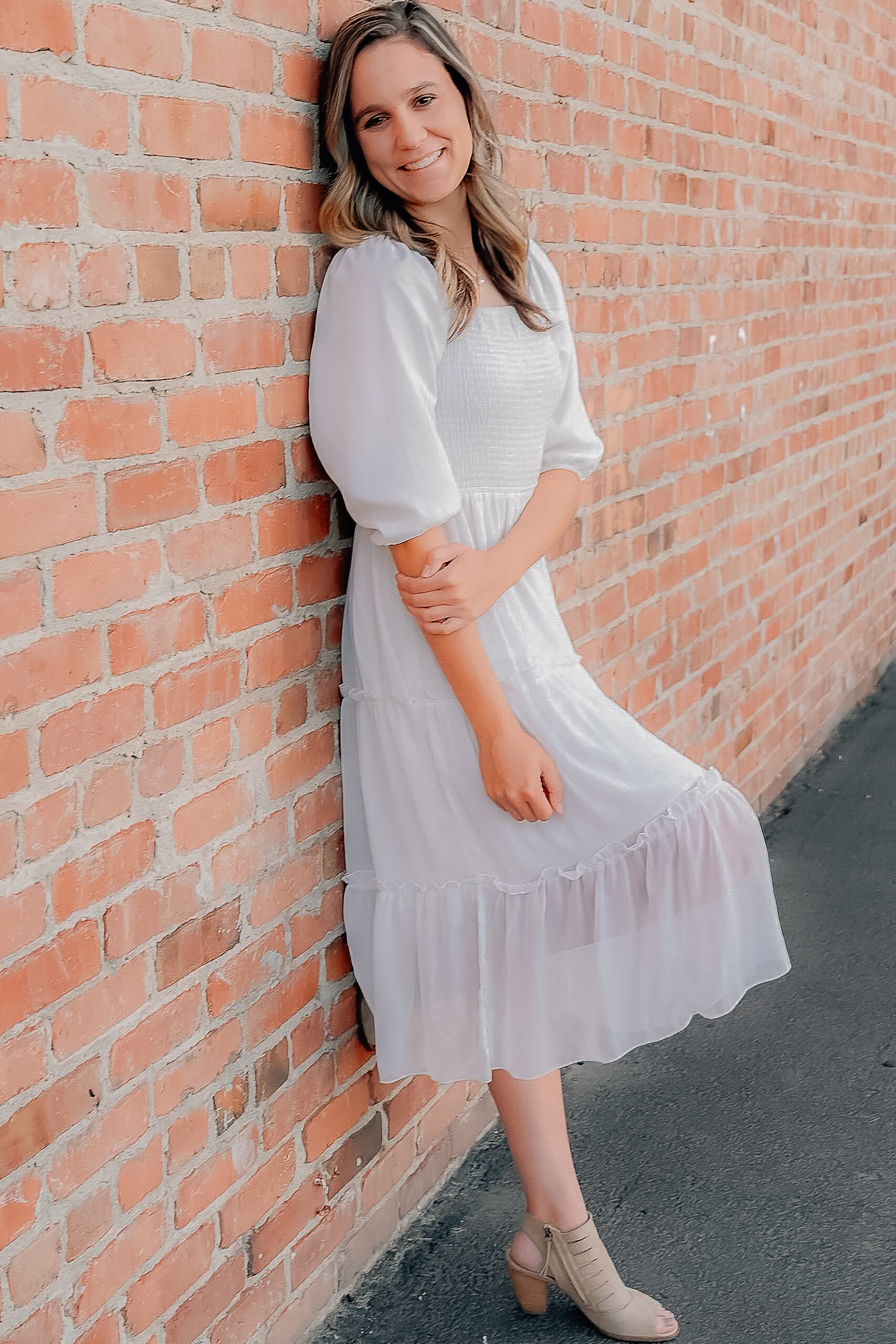 A senior girl wearing a white dress while leaning up against a brick wall. Her hand is clasping onto her elbow while leaving her other hand loose and relaxed.