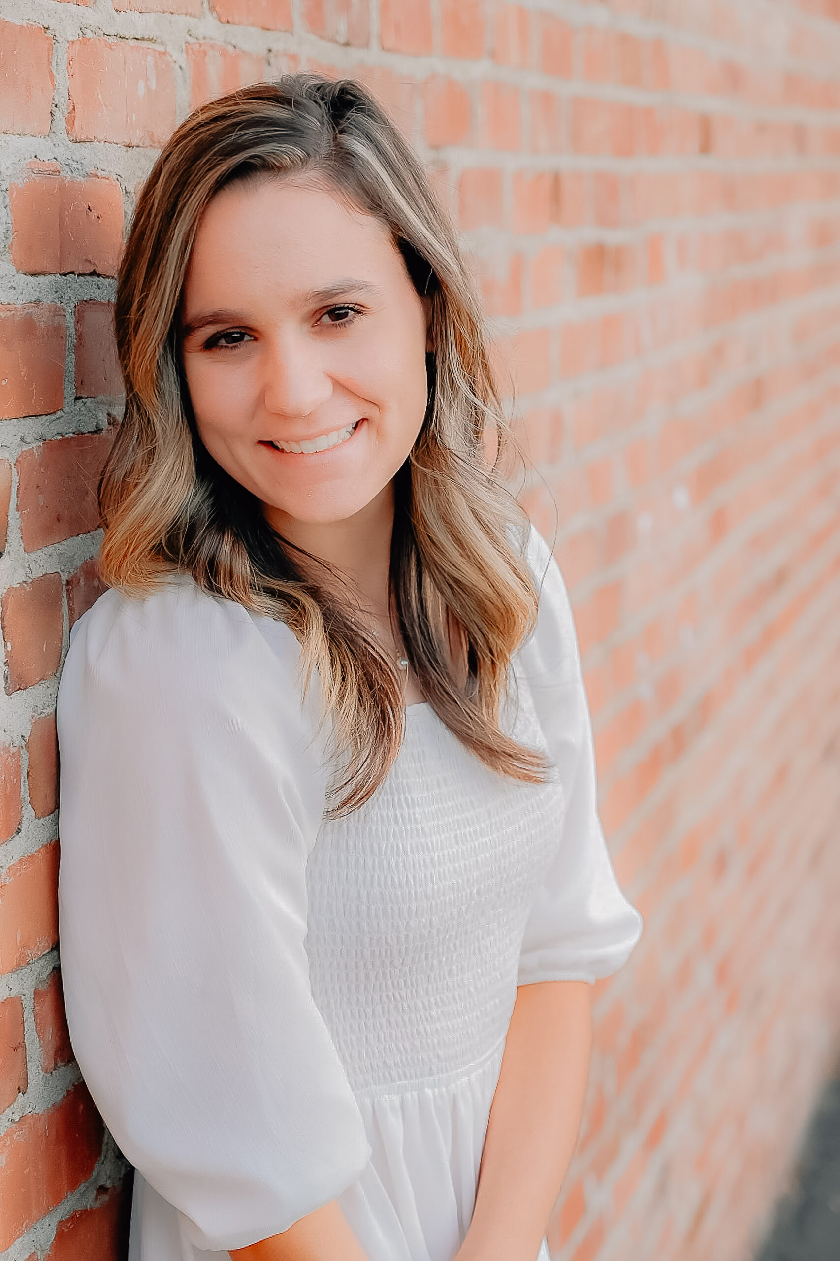 A senior girl wearing a white dress while leaning up against a brick wall. Her hand is clasping onto her wrist while leaving her other hand loose and relaxed.