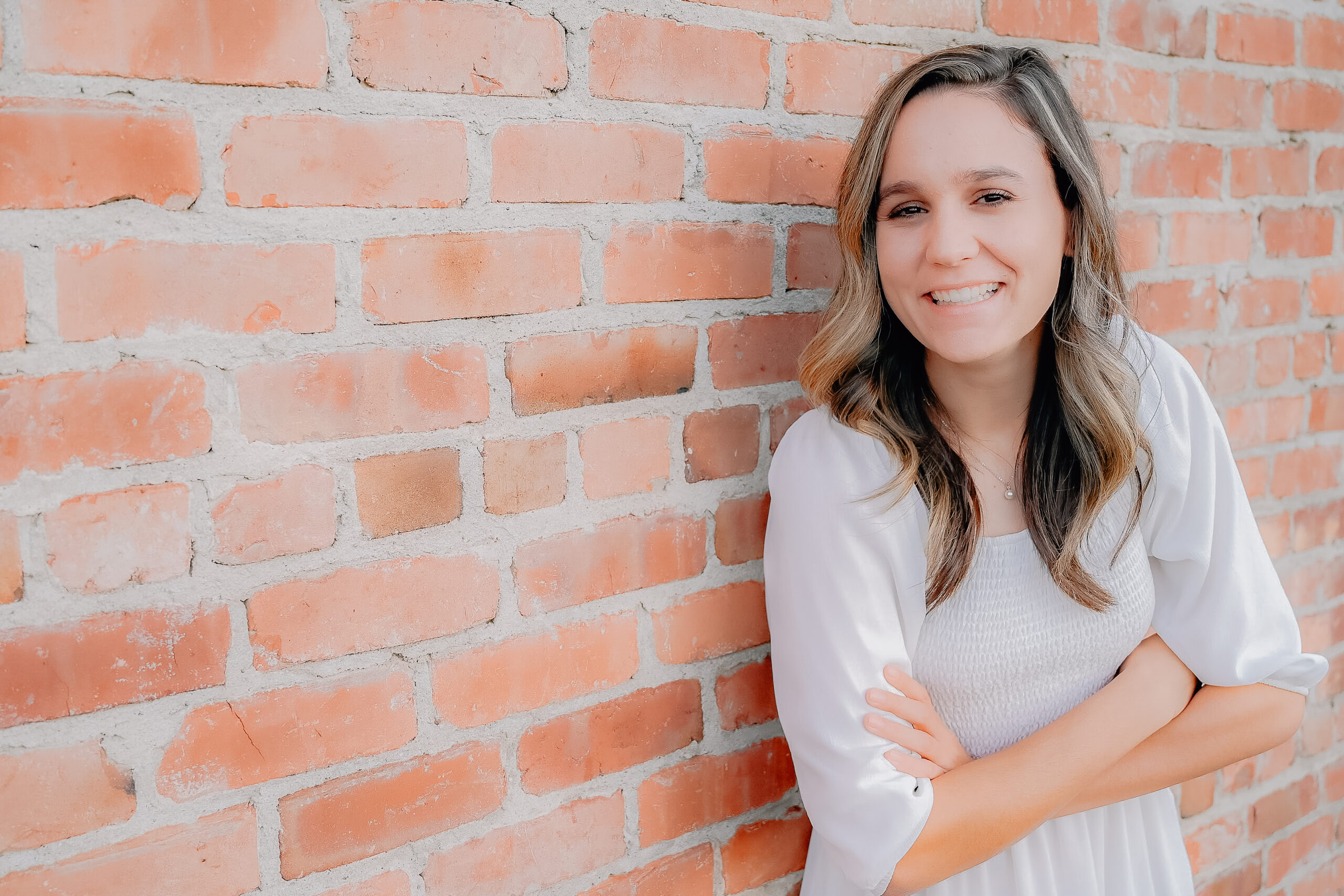 A senior girl wearing a white dress while leaning up against a brick wall folding her arms in front of her and laughing towards the camera.
