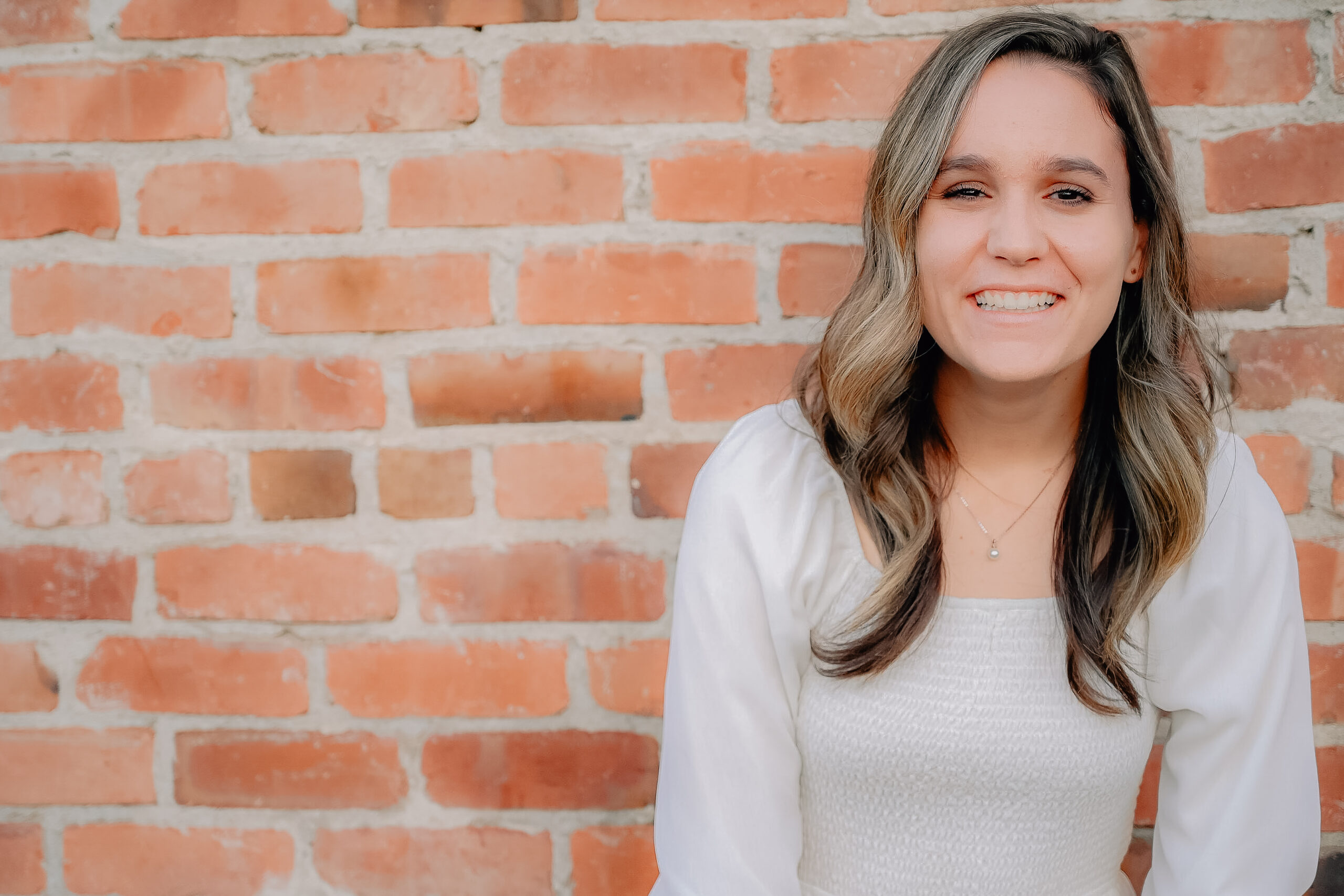 A senior girl wearing a white dress while leaning up against a brick wall and laughing towards the camera.